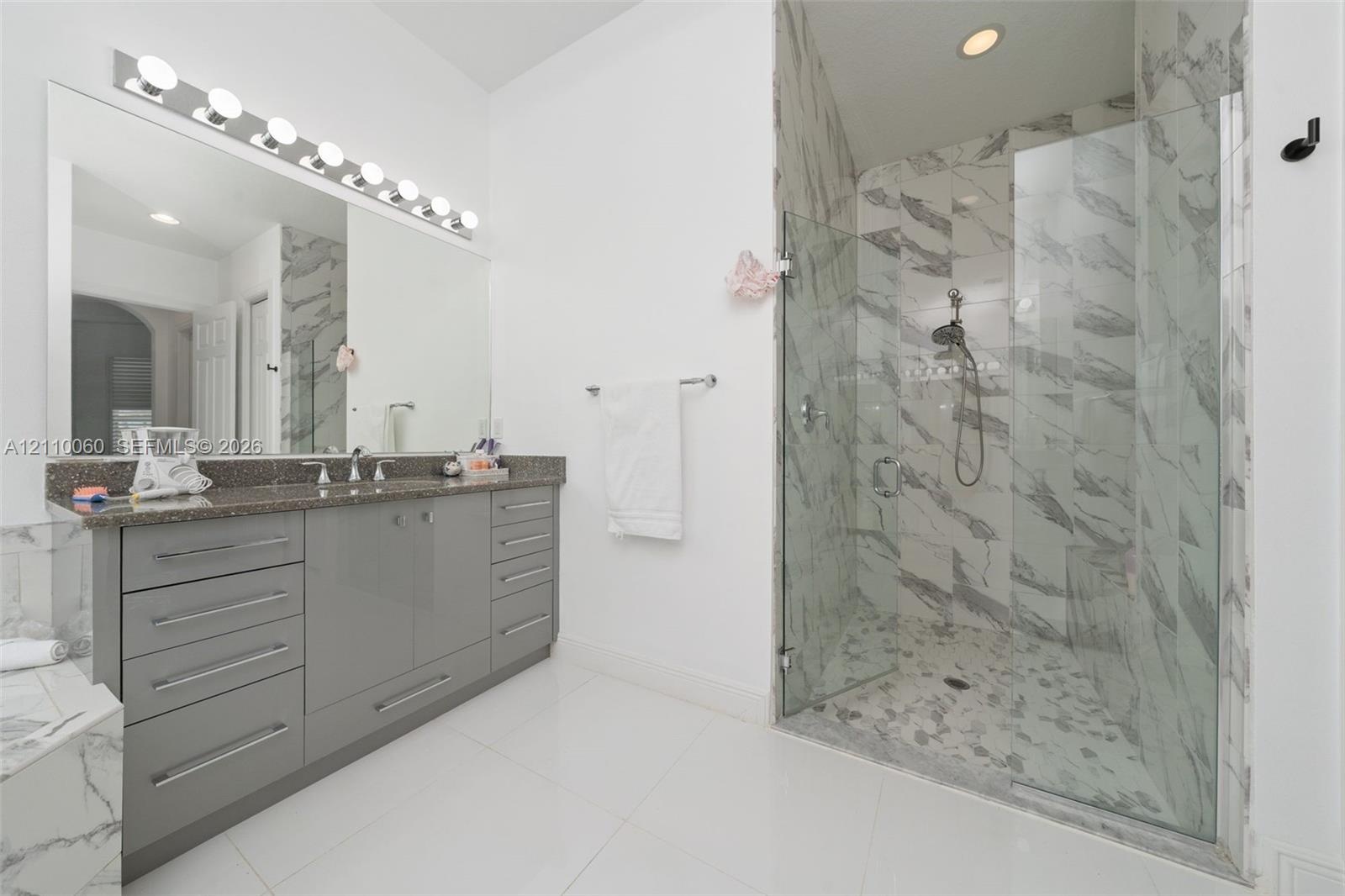 Bathroom featuring a dual-sink vanity with grey cabinetry, a solid-surface countertop, full-width mirror with multi-bulb lighting, and a glass-enclose