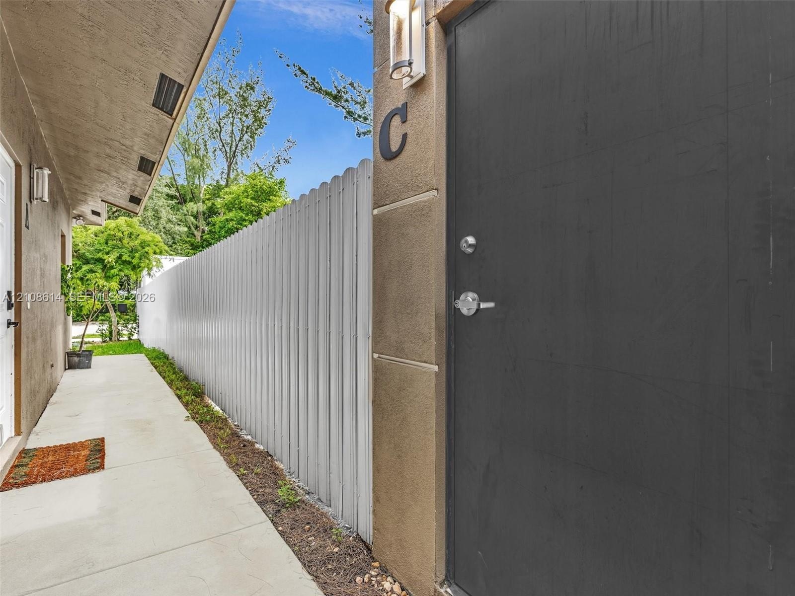 Exterior walkway featuring concrete paving, a white metal privacy fence, stucco siding, and a dark-toned solid entry door with integrated wall-mounted