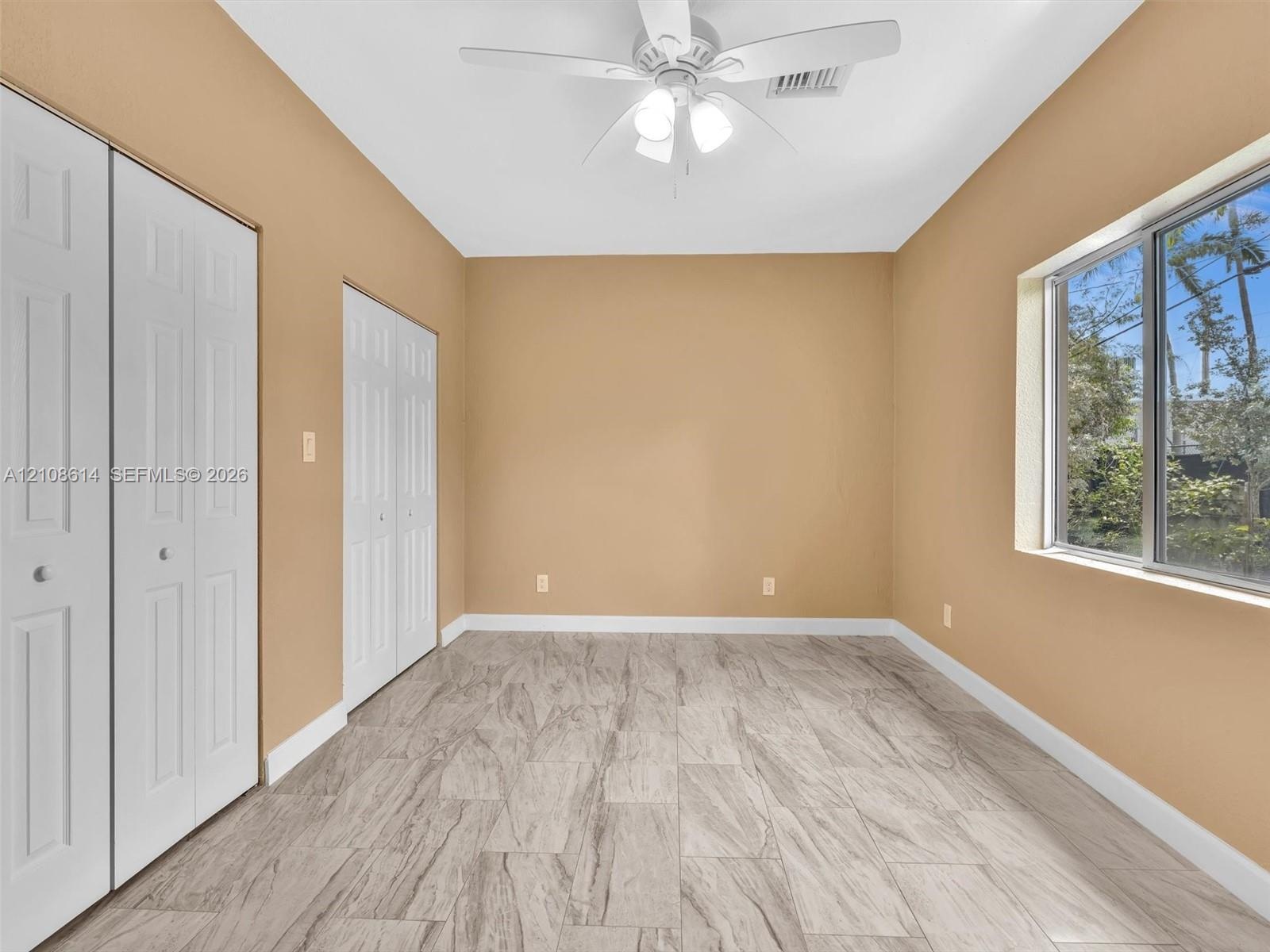 Neutral-toned room featuring stone-finish tile flooring, dual sets of bi-fold closet doors, and a ceiling-mounted light fixture with integrated fan