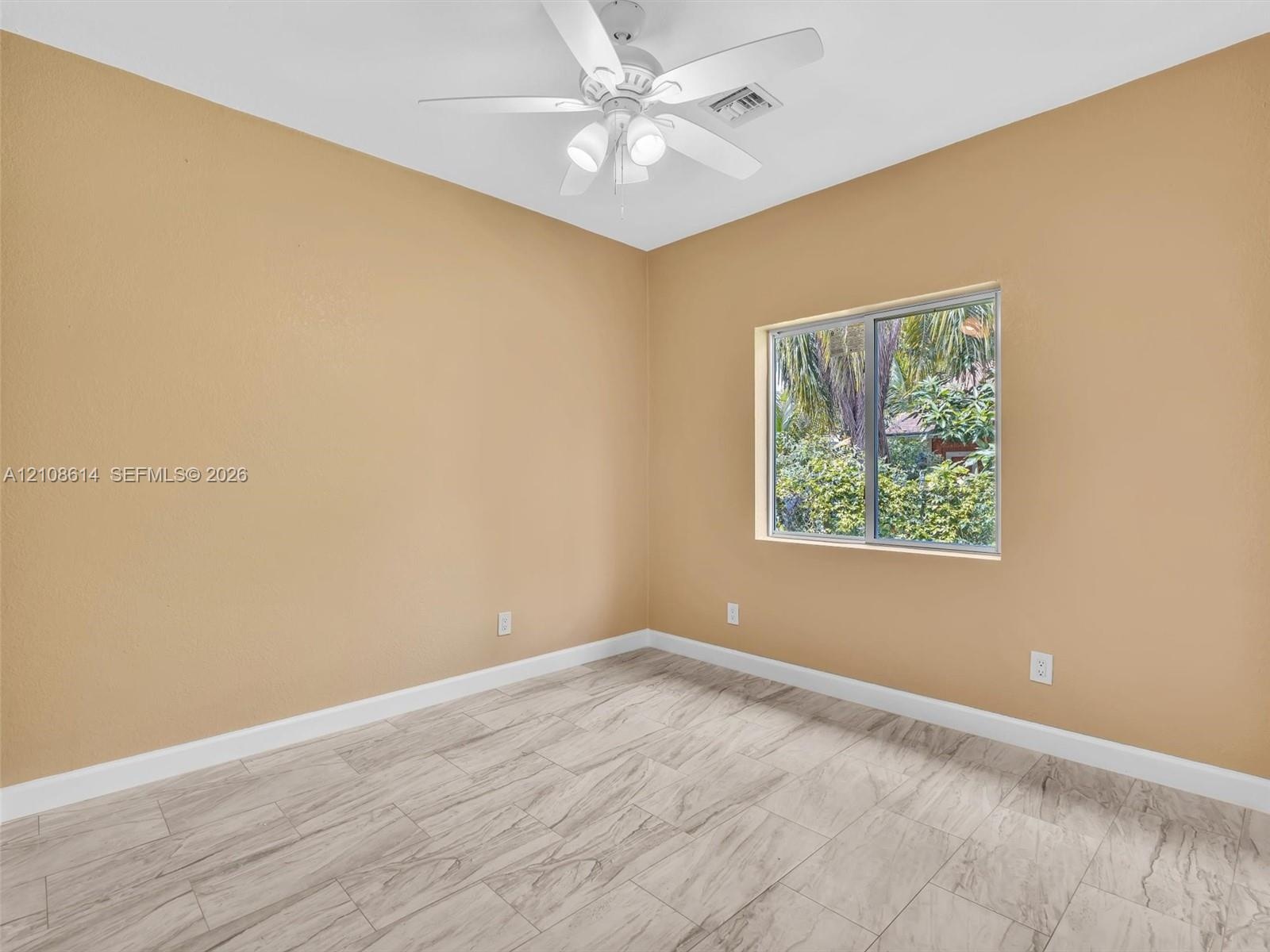 Neutral-toned interior space featuring wood-finish flooring, a white ceiling fan with integrated lighting, and a window with exterior greenery