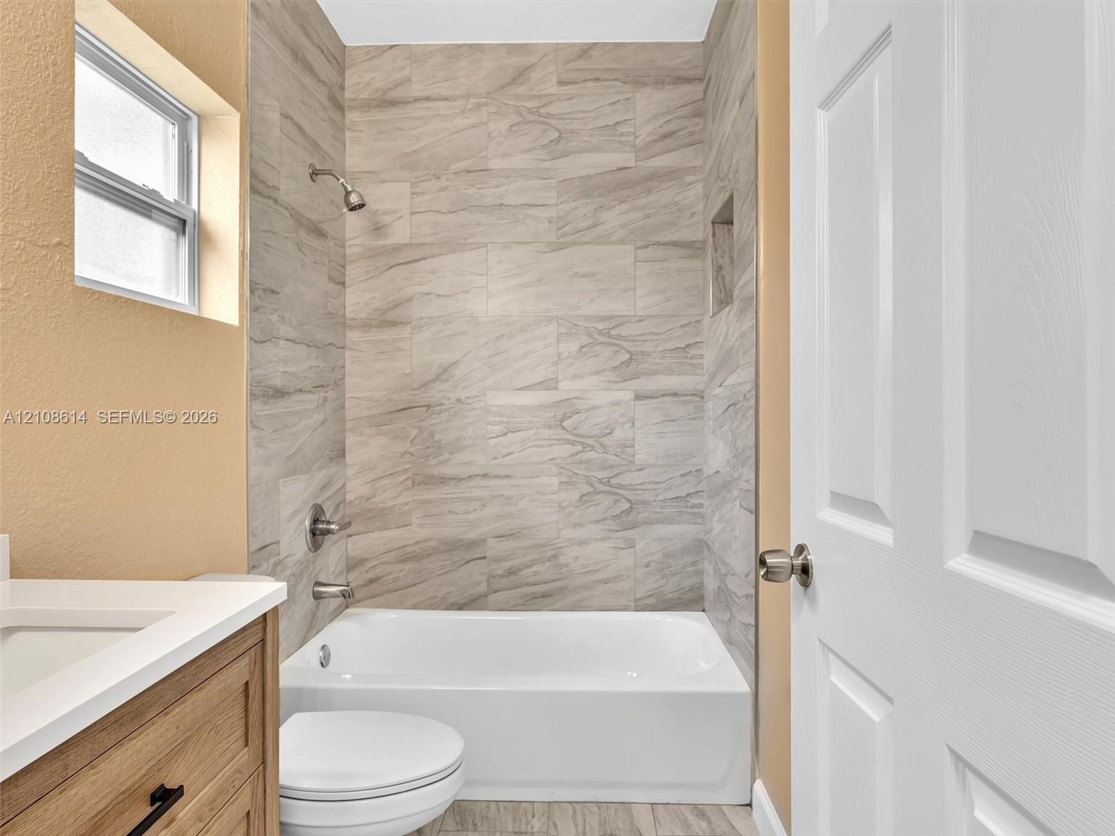 Bathroom featuring a tub-shower combination with floor-to-ceiling stone-look tile surround, built-in wall niche, wood-finish vanity with a rectangular
