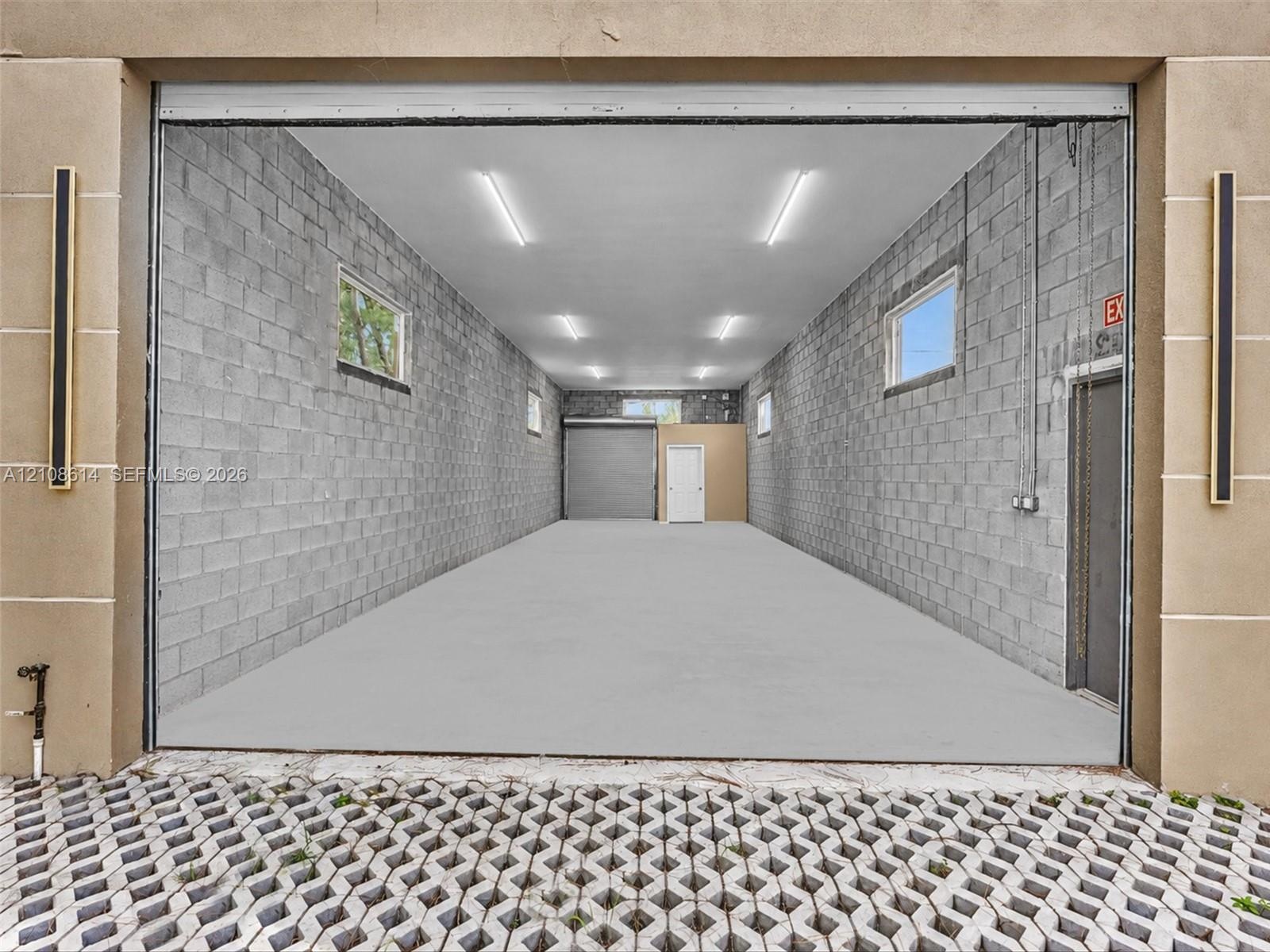 Expansive garage featuring concrete block walls, overhead lighting, multiple windows, a secondary roll-up door, and an exterior permeable paver apron