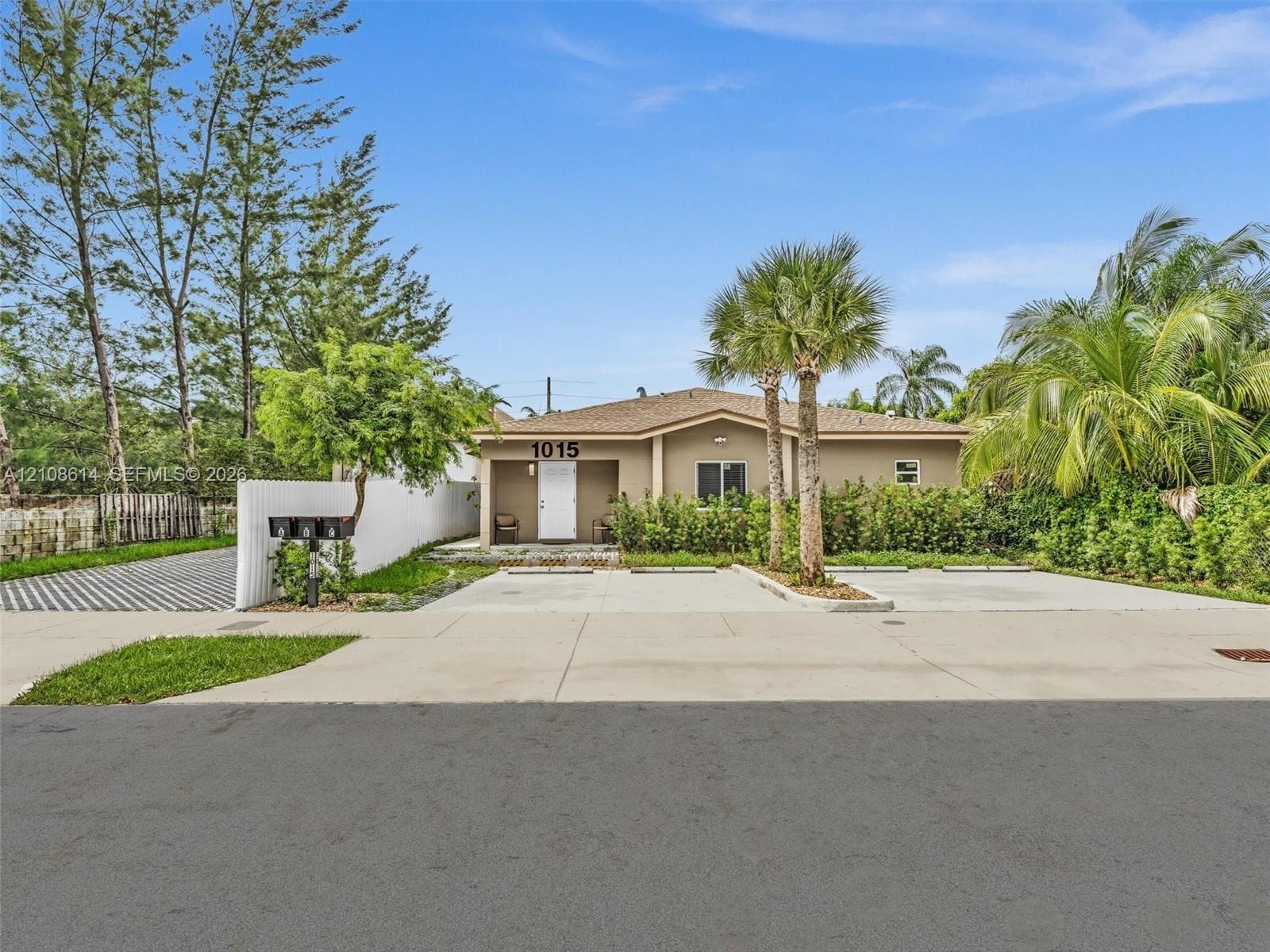 Single-story exterior featuring a stucco facade, covered entryway, concrete driveway, white vinyl fencing, and mature palm landscaping