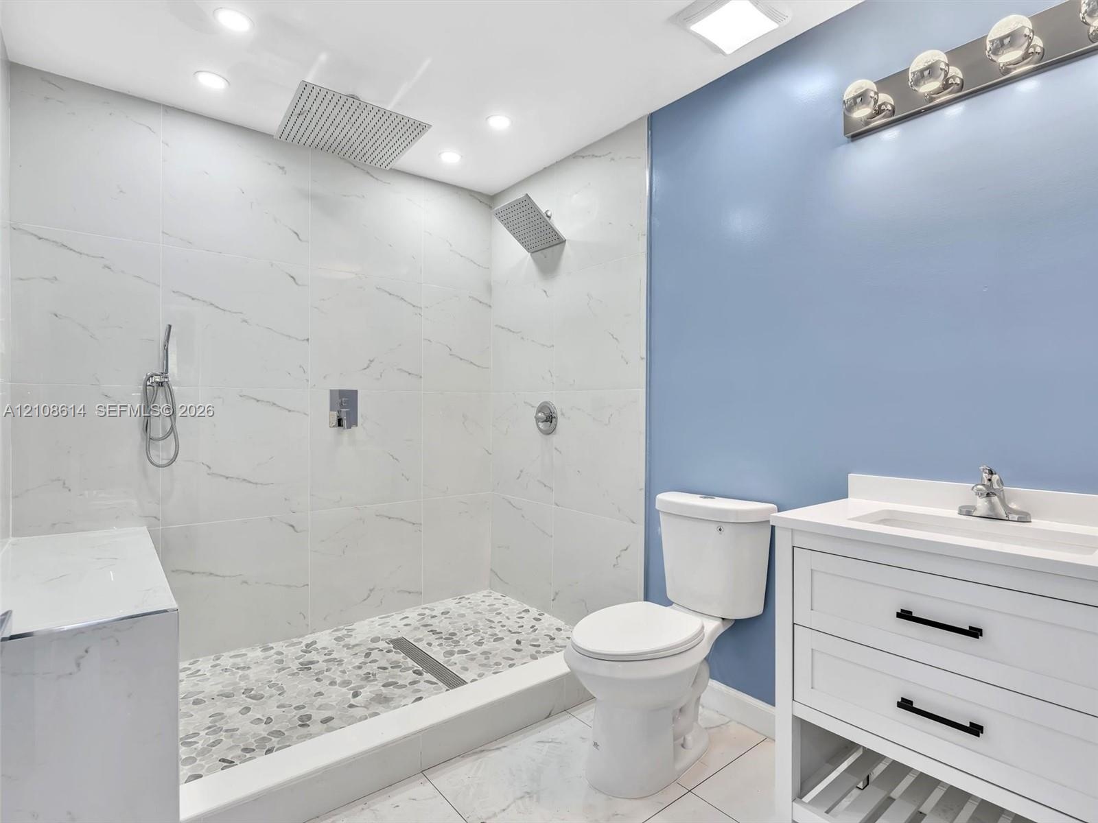 Walk-in shower featuring large-format tile walls, a pebble-stone floor, and a rainfall showerhead, complemented by a white vanity with integrated basi
