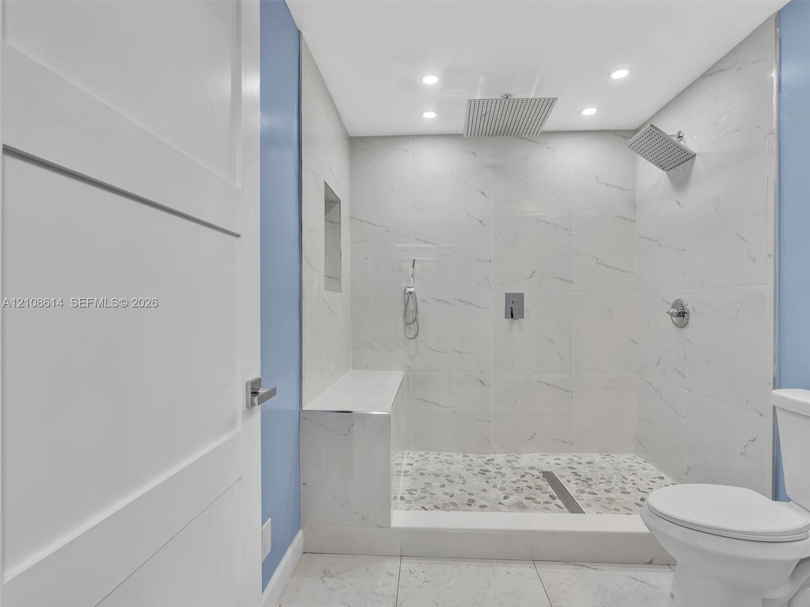 Walk-in shower featuring floor-to-ceiling stone-finish tile, a built-in stone bench, a recessed wall niche, a rain-style showerhead with handheld spra