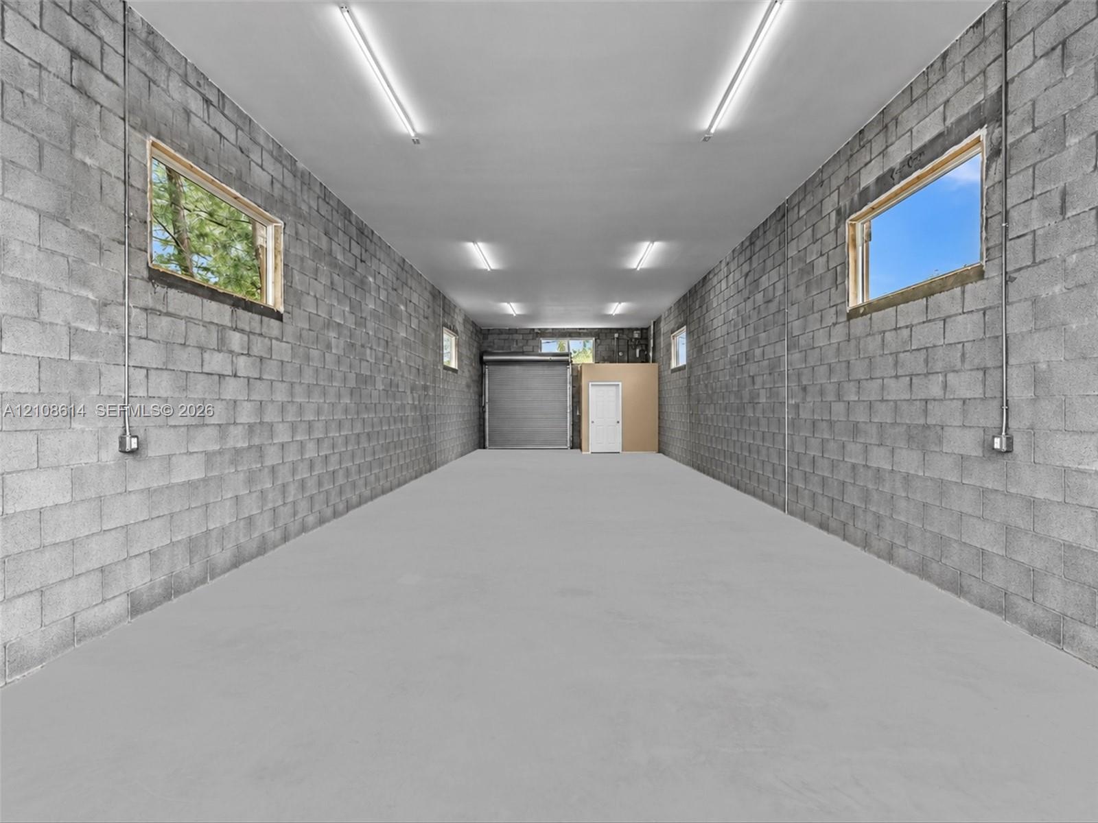 Expansive workspace featuring concrete block walls, a smooth concrete floor, multiple windows, overhead lighting, and a metal roll-up door
