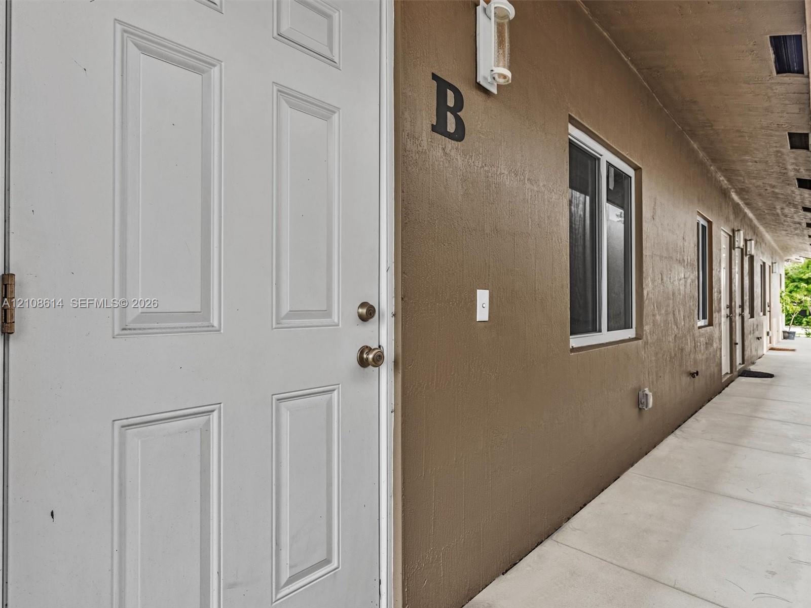 Exterior entrance featuring a six-panel white door, smooth stucco finish, wall-mounted light fixture, and concrete walkway