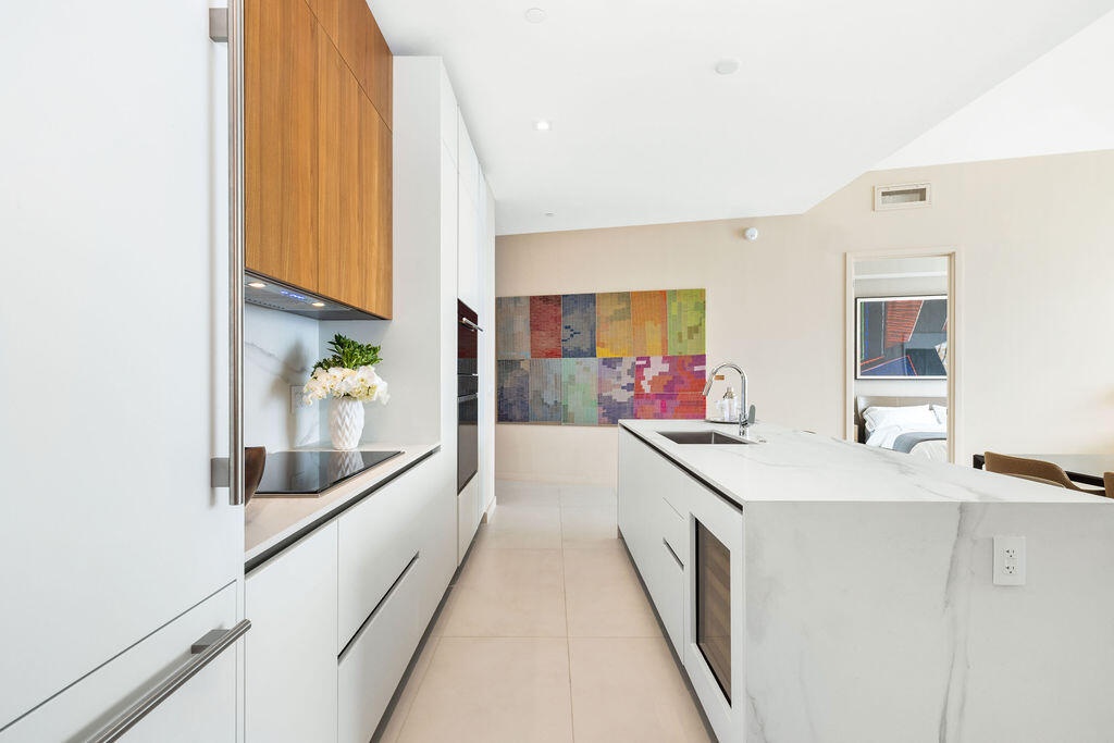 Kitchen featuring white flat-panel cabinetry, wood-finish upper cabinets, stone-surfaced waterfall island, built-in wall oven, and integrated wine coo