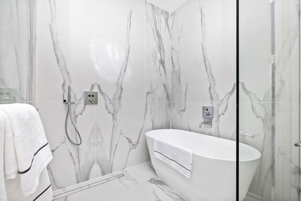 Freestanding soaking tub featuring a wall-mounted faucet, floor-to-ceiling large-format stone-finish tile surround, handheld shower fixture, and glass