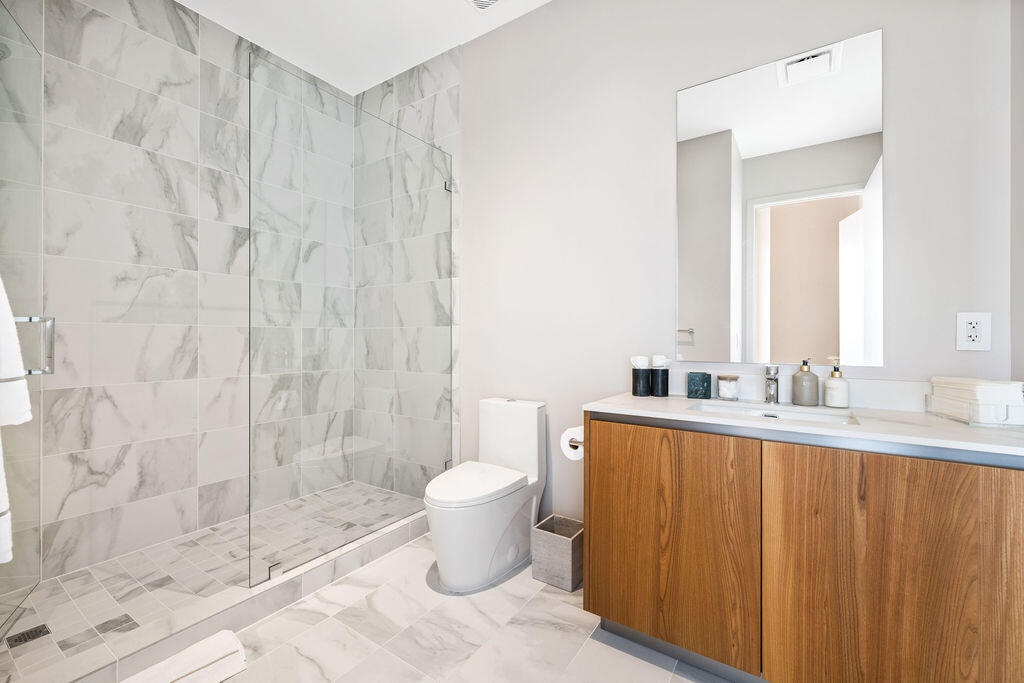 Bathroom featuring a walk-in shower with frameless glass enclosure, floor-to-ceiling stone-pattern tile, wood-finish cabinetry, white countertop, and 