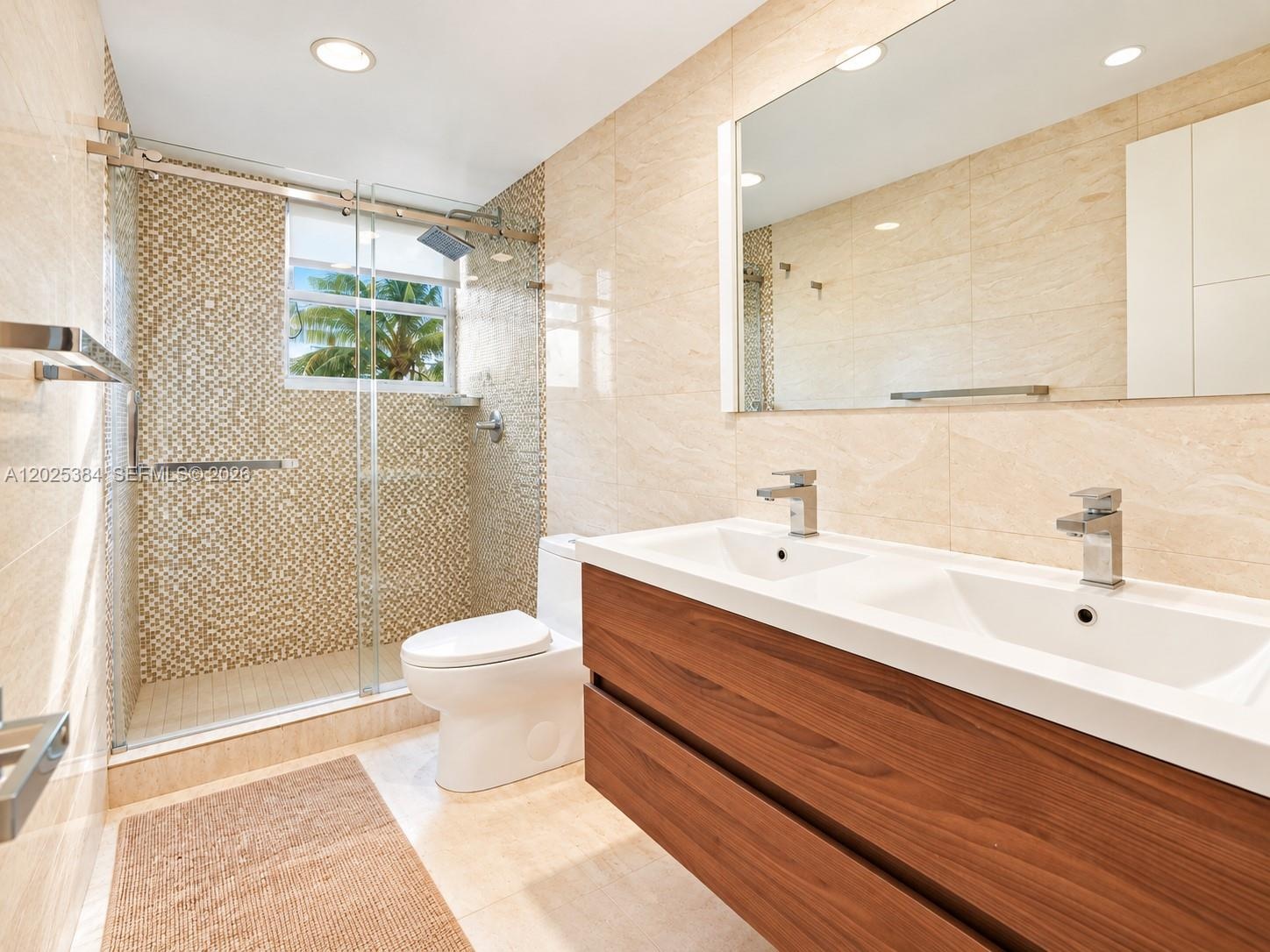 Double vanity with a wood-finish cabinet and integrated dual sinks, walk-in shower featuring glass doors and mosaic tile accents, wall-to-wall mirror,