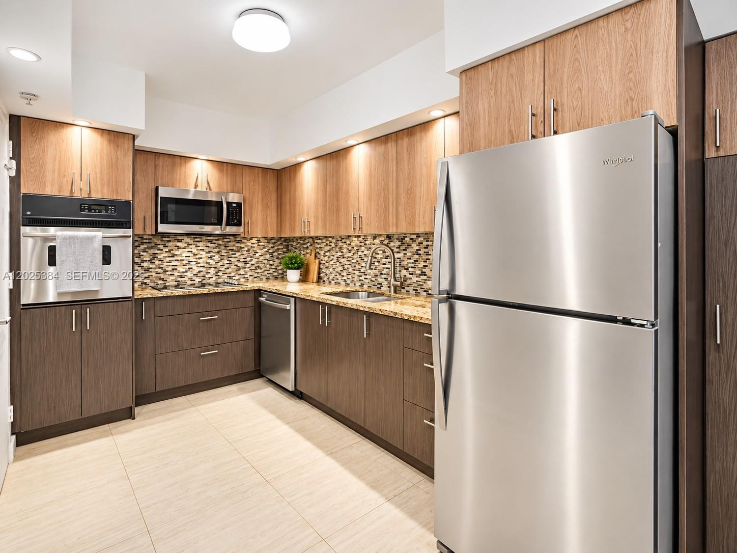 Kitchen featuring two-tone flat-panel cabinetry, stone-finish countertops, multi-tonal glass tile backsplash, stainless steel appliances, and large-fo