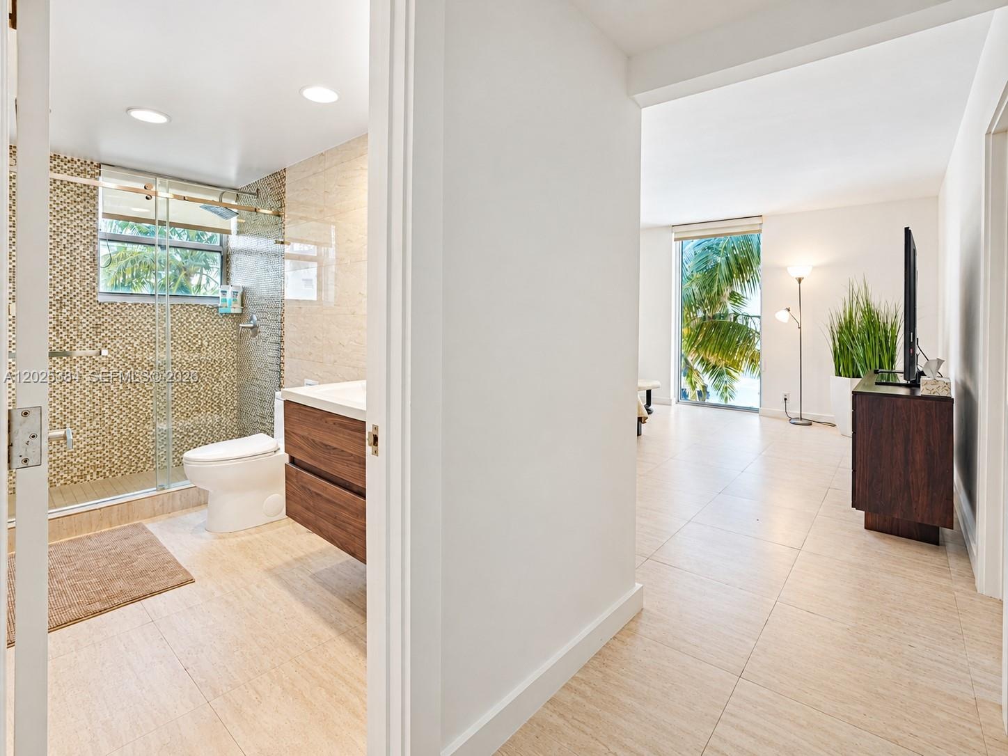 Bathroom featuring a wood-finish floating vanity, glass-enclosed shower with tiled walls, and large-format tile flooring extending into the living are