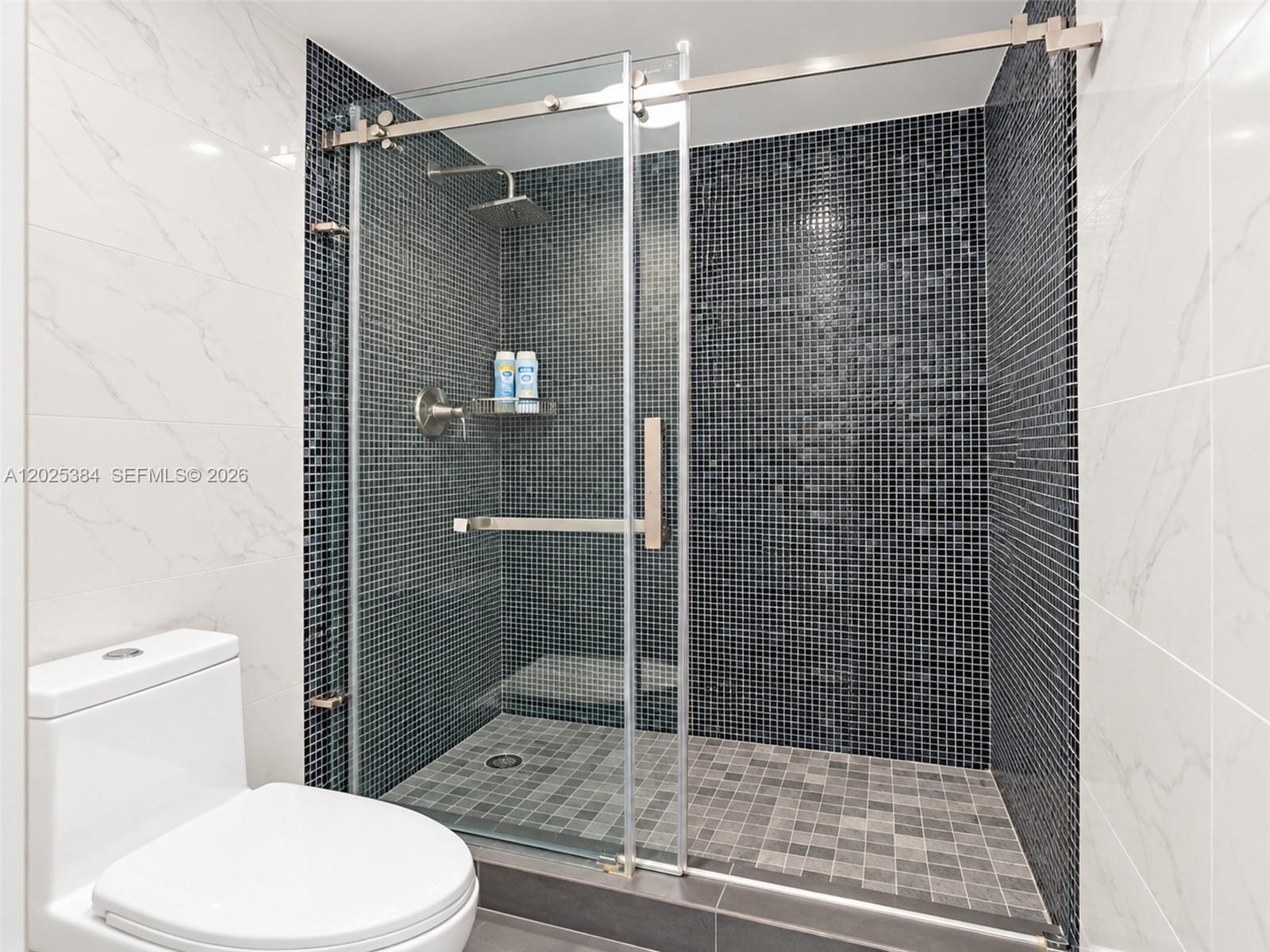 Walk-in shower featuring a sliding glass door, high-contrast black mosaic tile surround, built-in recessed shelving, stainless steel rainfall shower h