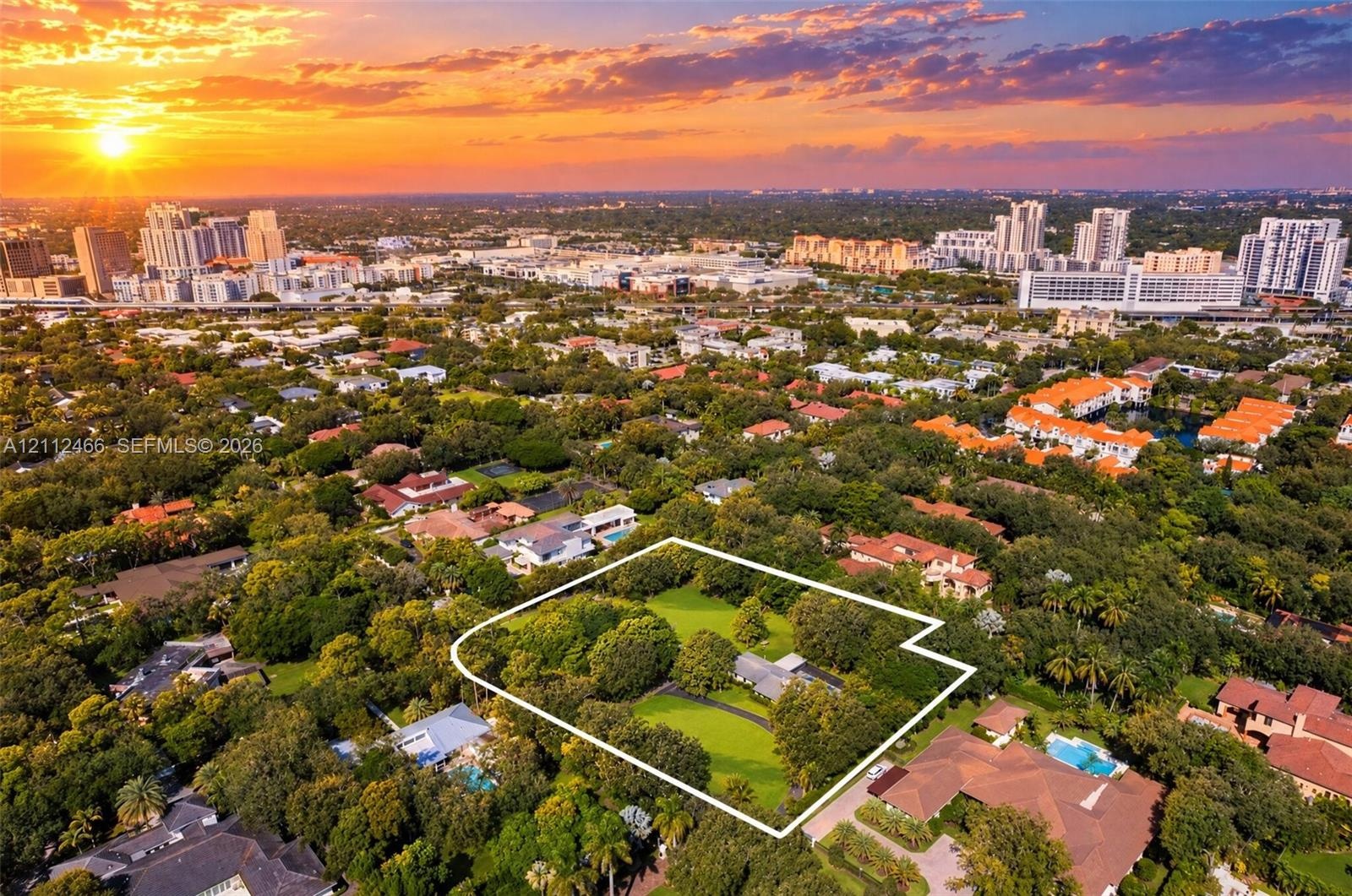 Aerial perspective of a sprawling property featuring mature trees, expansive manicured lawn, paved driveway, and prominent cityscape backdrop