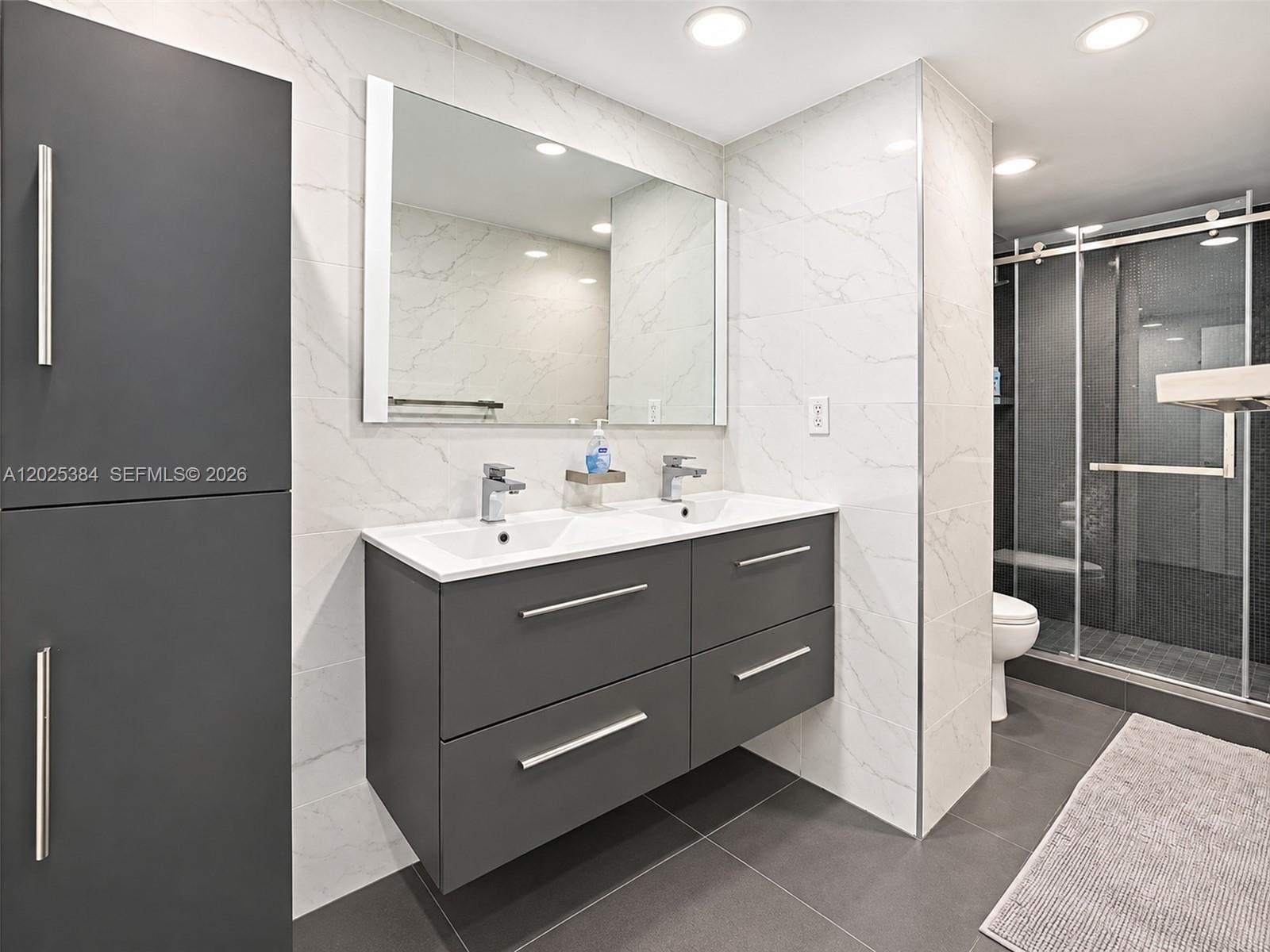 Floating double vanity with a quartz countertop, rectangular vessel sinks, floor-to-ceiling large-format tile wall cladding, and a glass-enclosed show