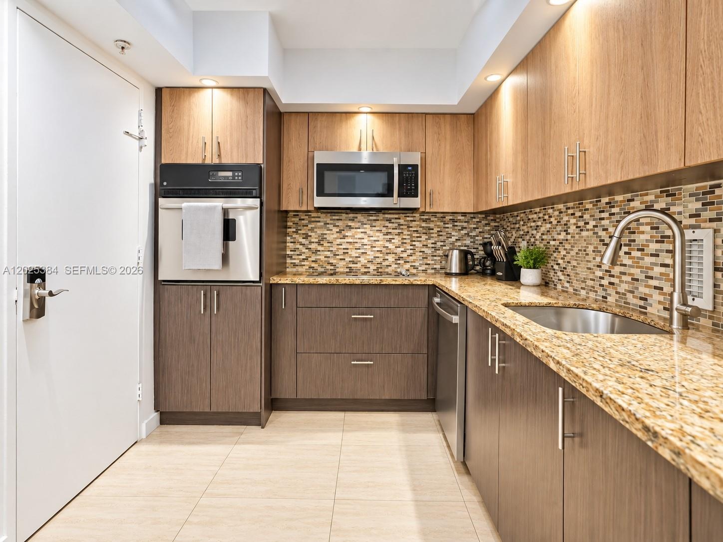 Kitchen featuring dual-toned flat-panel cabinetry, light-toned stone countertops, a mosaic tile backsplash, stainless steel appliances, and tile floor