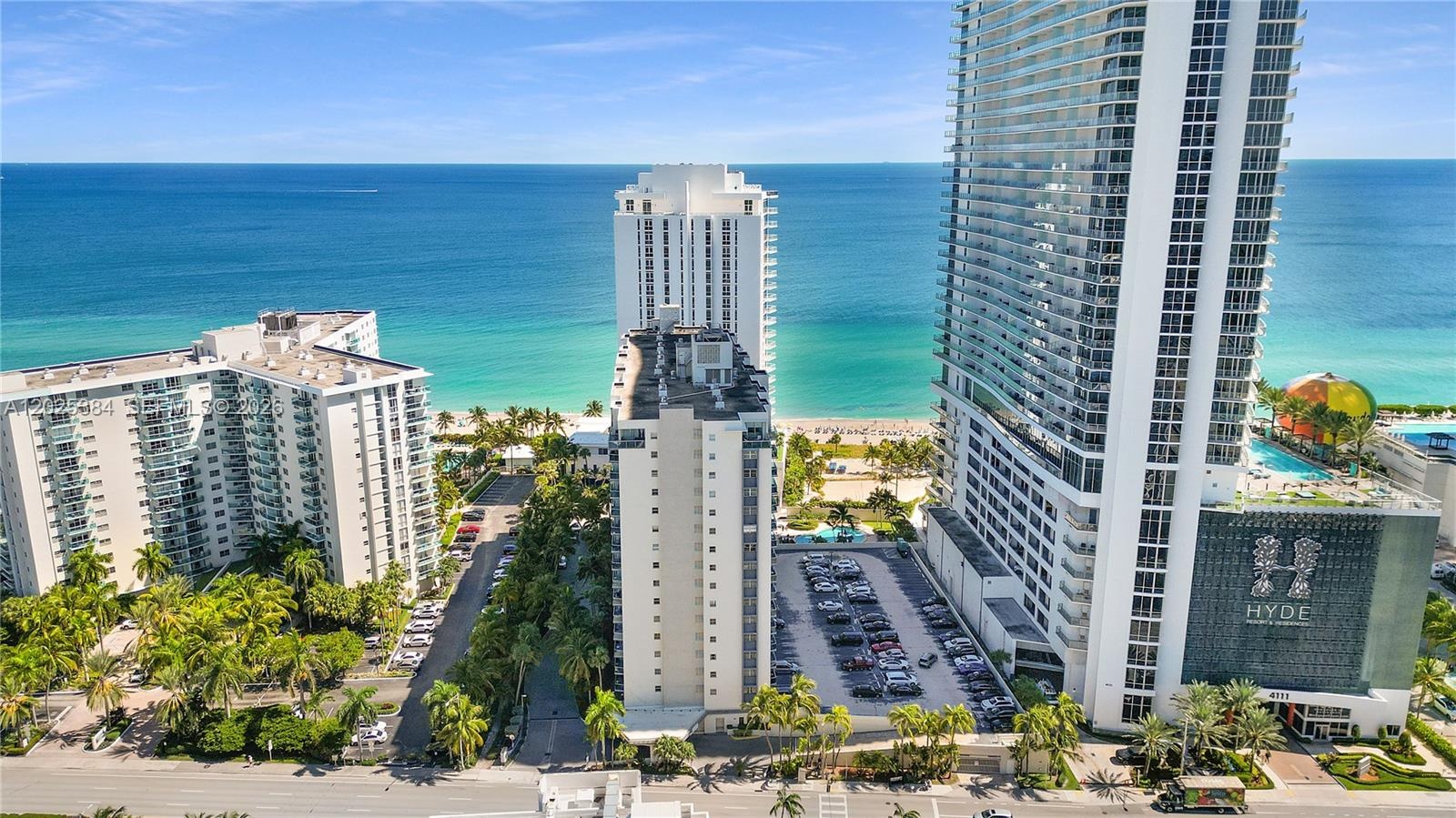Aerial perspective of a multi-structure beachfront complex featuring high-rise architecture, paved surface parking, and direct access to the coastline