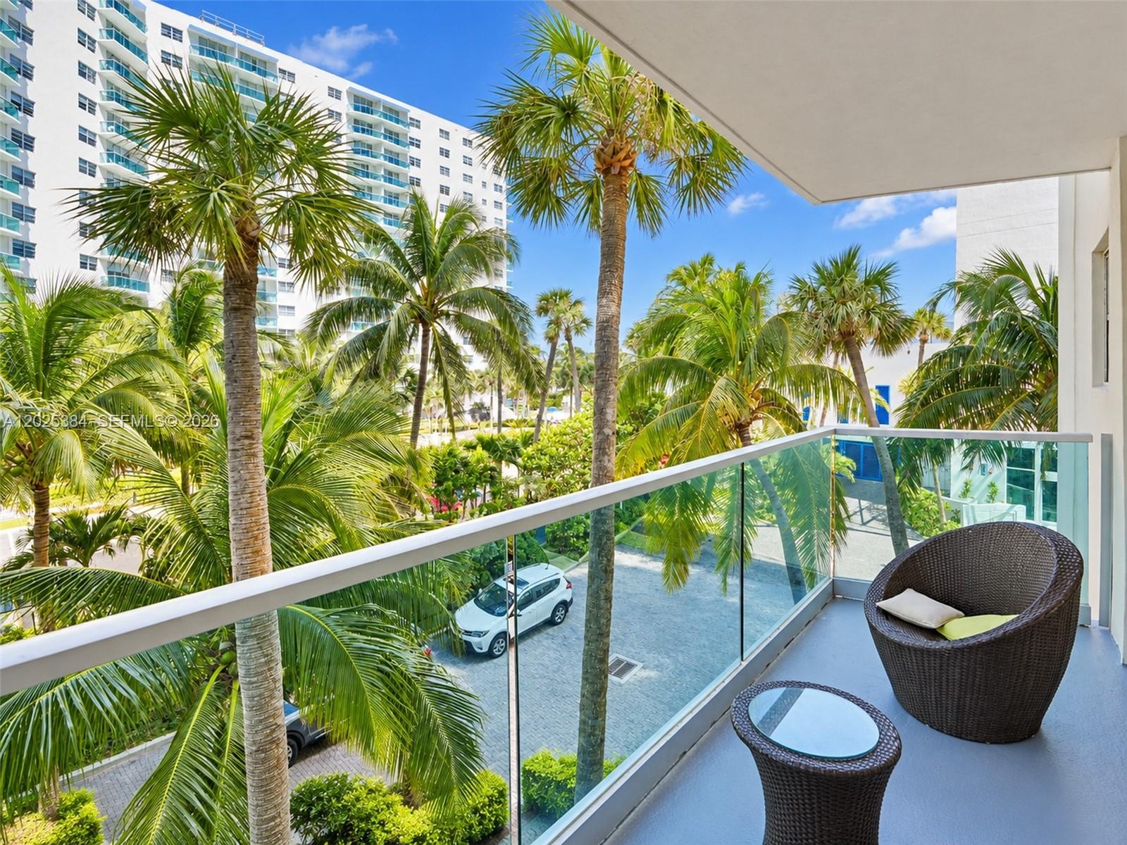 Covered balcony featuring glass railing panels and metal handrails, framed by mature palm trees