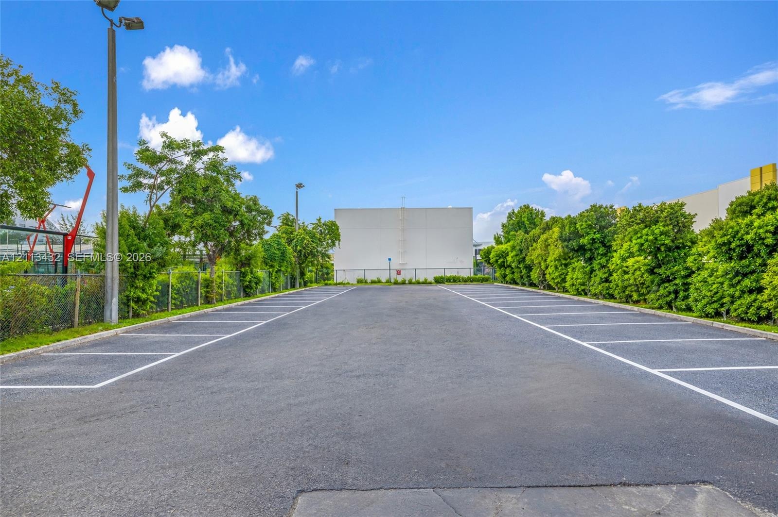 Asphalt parking area featuring marked spaces, perimeter chain-link fencing, mature trees, and installed exterior lighting poles