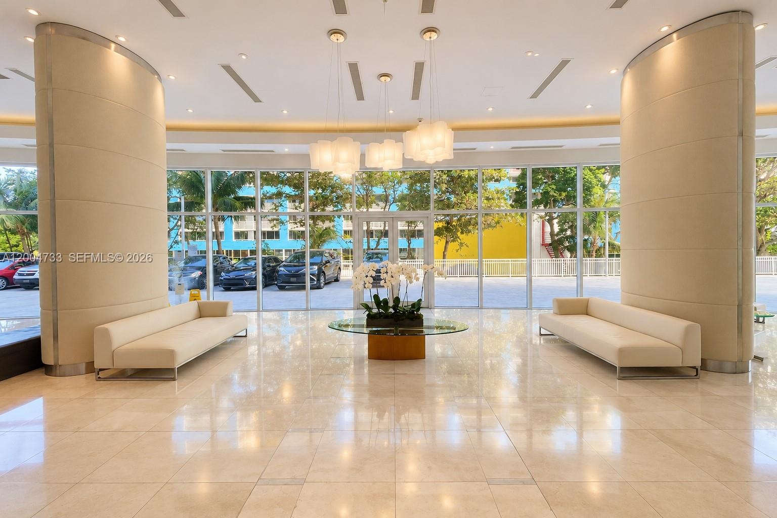Grand lobby featuring polished stone flooring, expansive floor-to-ceiling glass paneling, dual curved structural columns, and contemporary suspended m