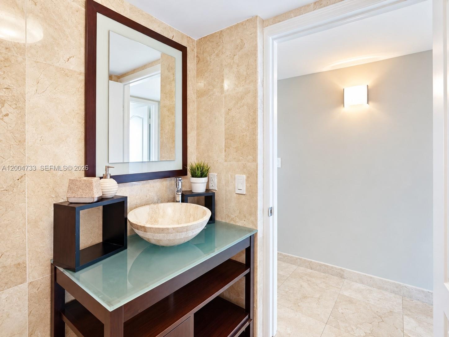 Bathroom featuring stone wall and floor tile, a dark wood-finish vanity with a glass countertop, a stone vessel sink, and a rectangular wall mirror wi