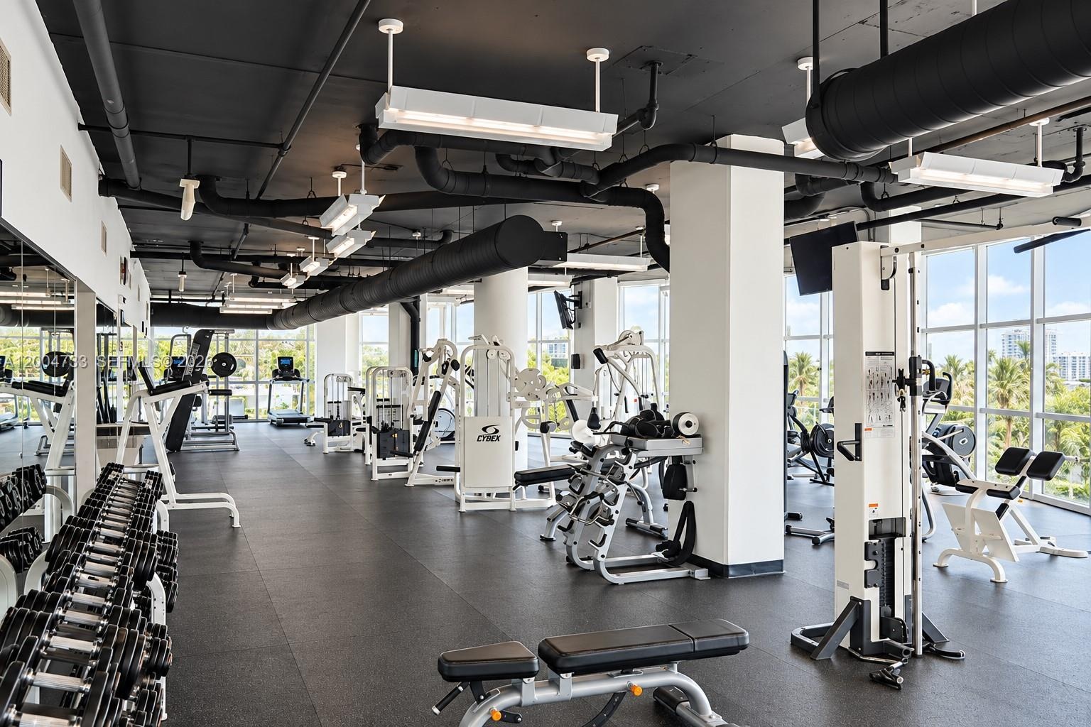 Fitness center featuring industrial-style exposed ceiling, rubberized flooring, expansive floor-to-ceiling windows, and wall-to-wall mirrors