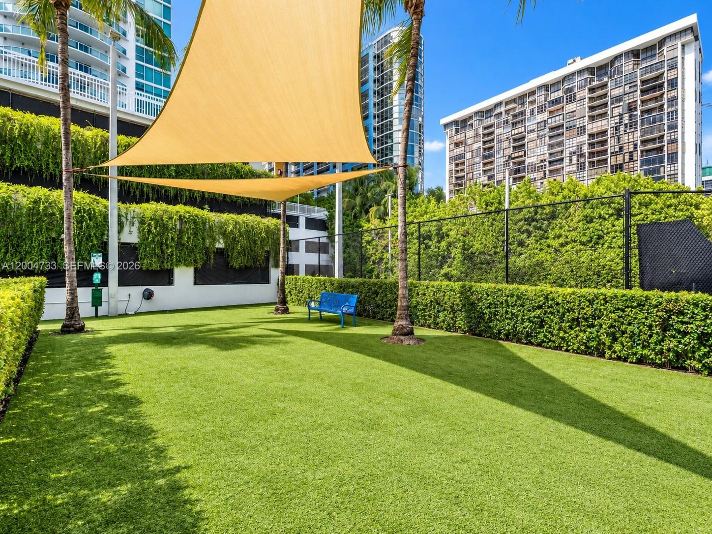Outdoor recreational space featuring synthetic turf, perimeter privacy hedging, mature palm trees, and overhead shade sails