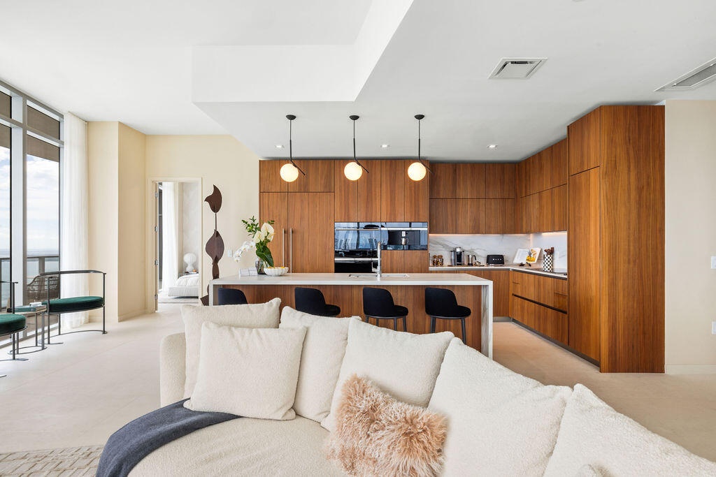 Modern kitchen featuring flat-panel wood cabinetry, a waterfall-edge kitchen island, stone backsplash, built-in wall oven, and three globe pendant lig