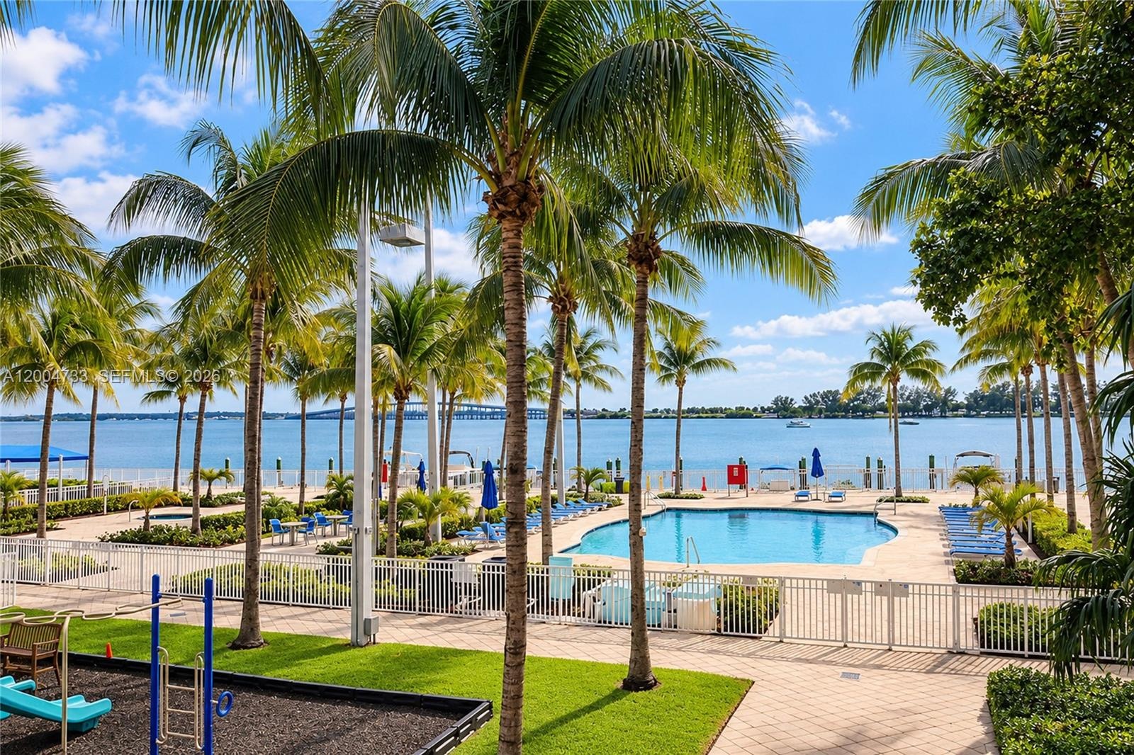 Community swimming pool with a paved deck, manicured green space, and mature palm trees, featuring a gated perimeter and water frontage