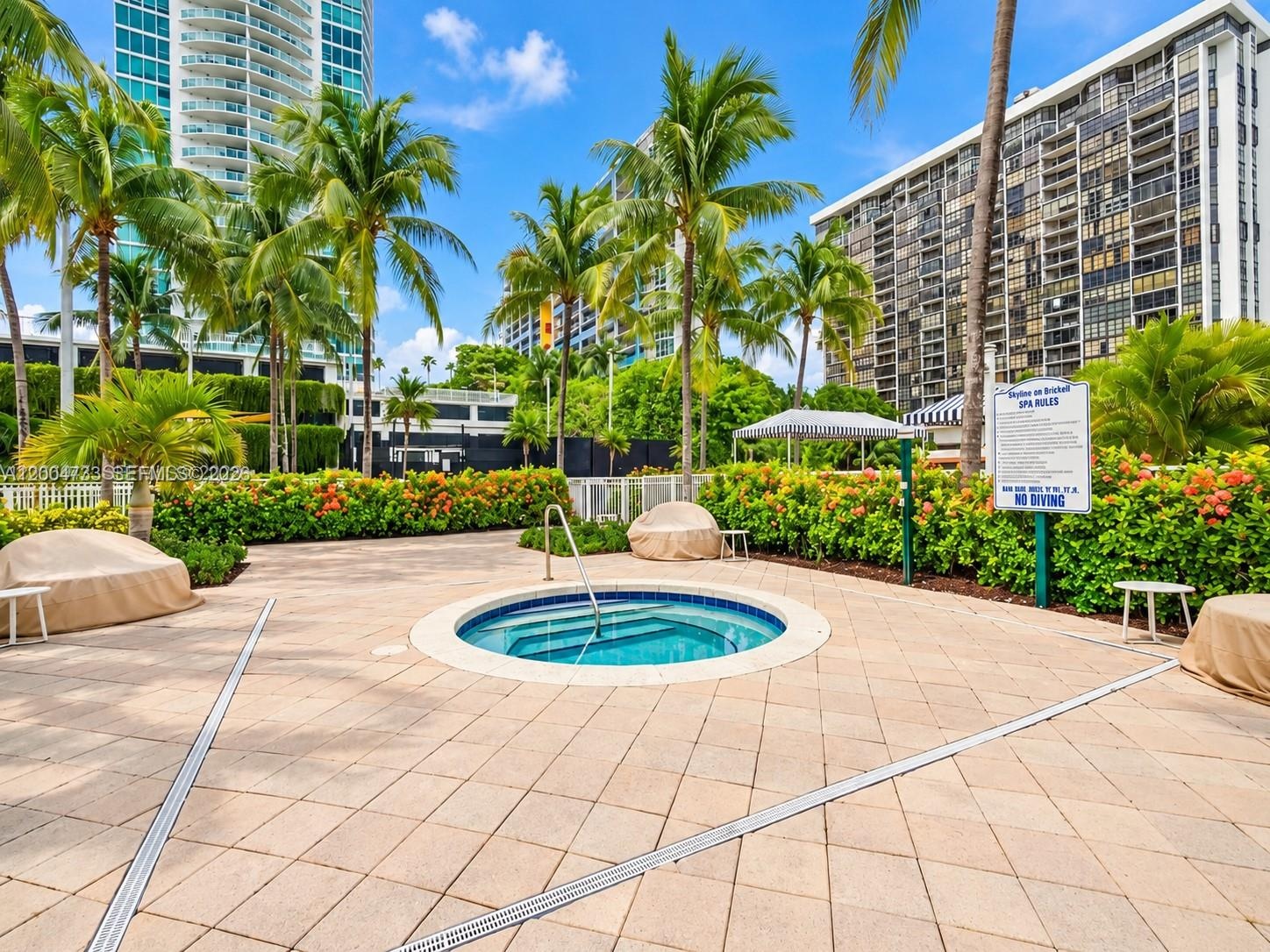 In-ground spa with stone tile surround, paver patio, metal handrail, perimeter hedges, and mature palms