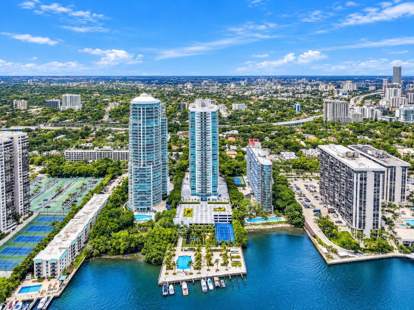 Aerial perspective featuring waterfront high-rise towers, a private marina, multiple swimming pools, and dedicated tennis courts