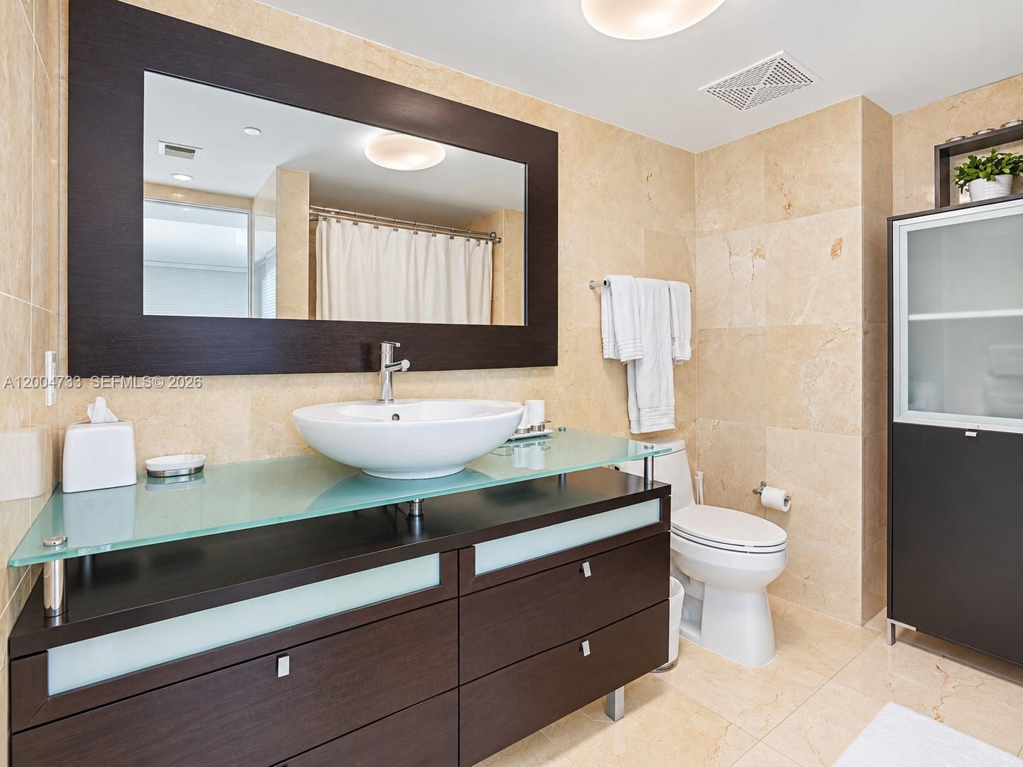 Bathroom featuring a vanity with a frosted glass countertop, vessel sink, dark wood-finish cabinetry, a large framed mirror, and natural stone tile fl