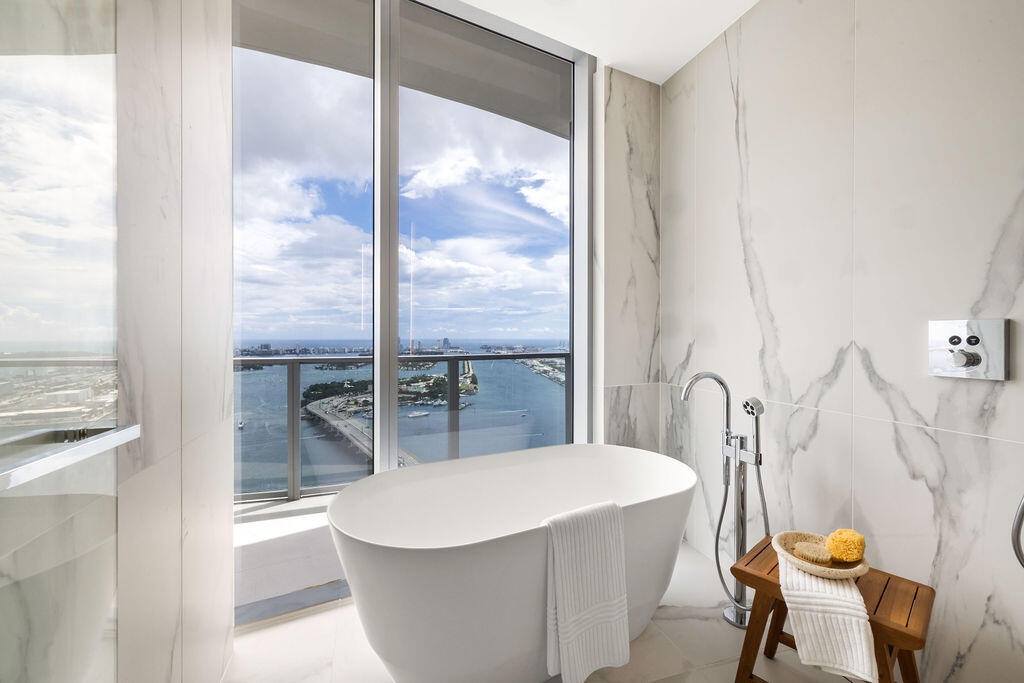 Freestanding soaking tub with polished chrome floor-mounted faucet, floor-to-ceiling porcelain wall tile, expansive window wall, and integrated balcon