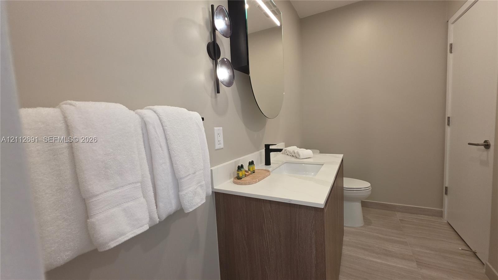 Modern vanity with wood-finish cabinetry, white countertop, integrated rectangular sink, matte black faucet, and oval mirror with wall-mounted lightin