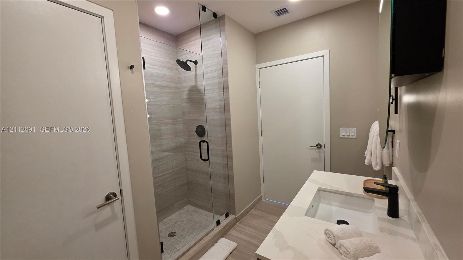 Bathroom featuring a walk-in shower with frameless glass enclosure, horizontal stone-finish tile surround, recessed lighting, white vanity with stone-