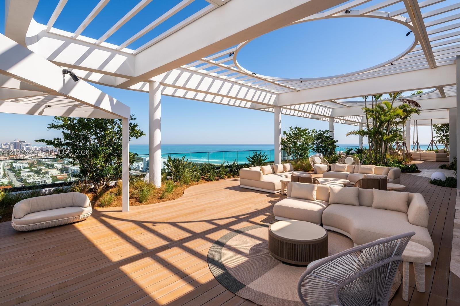 Expansive rooftop terrace featuring a white pergola structure with a circular cutout, wood-finish decking, glass panel railings, and perimeter landsca
