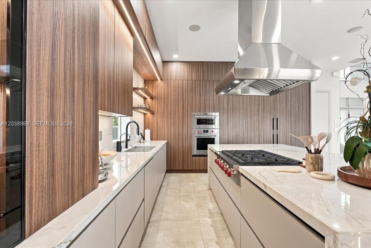 Kitchen featuring full-height wood-finish cabinetry, waterfall-edge stone countertops, stainless steel professional-grade range hood, built-in wall ov