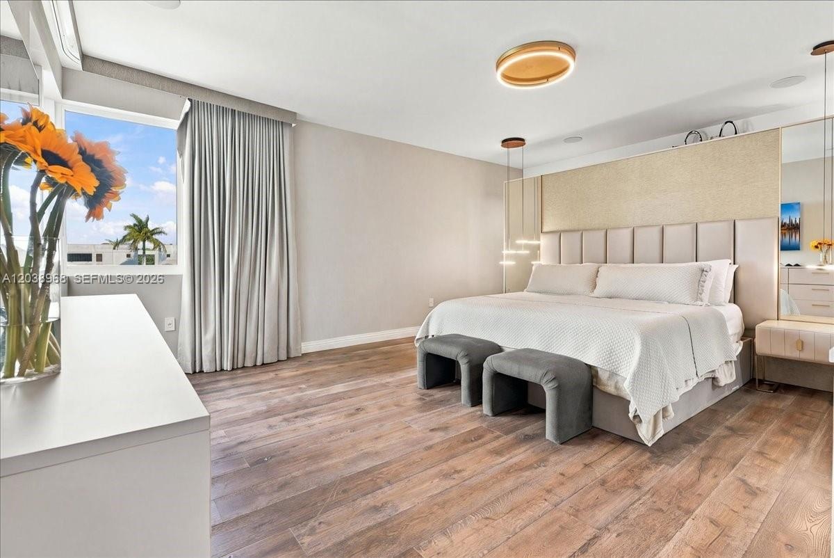Spacious room featuring wood-finish flooring, floor-to-ceiling drapery, a custom upholstered headboard wall, tiered pendant lighting, and a flush-moun