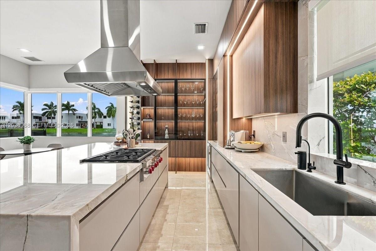 Contemporary kitchen featuring a stone-topped waterfall island, custom wood-finish cabinetry, stainless steel range hood, integrated display shelving 
