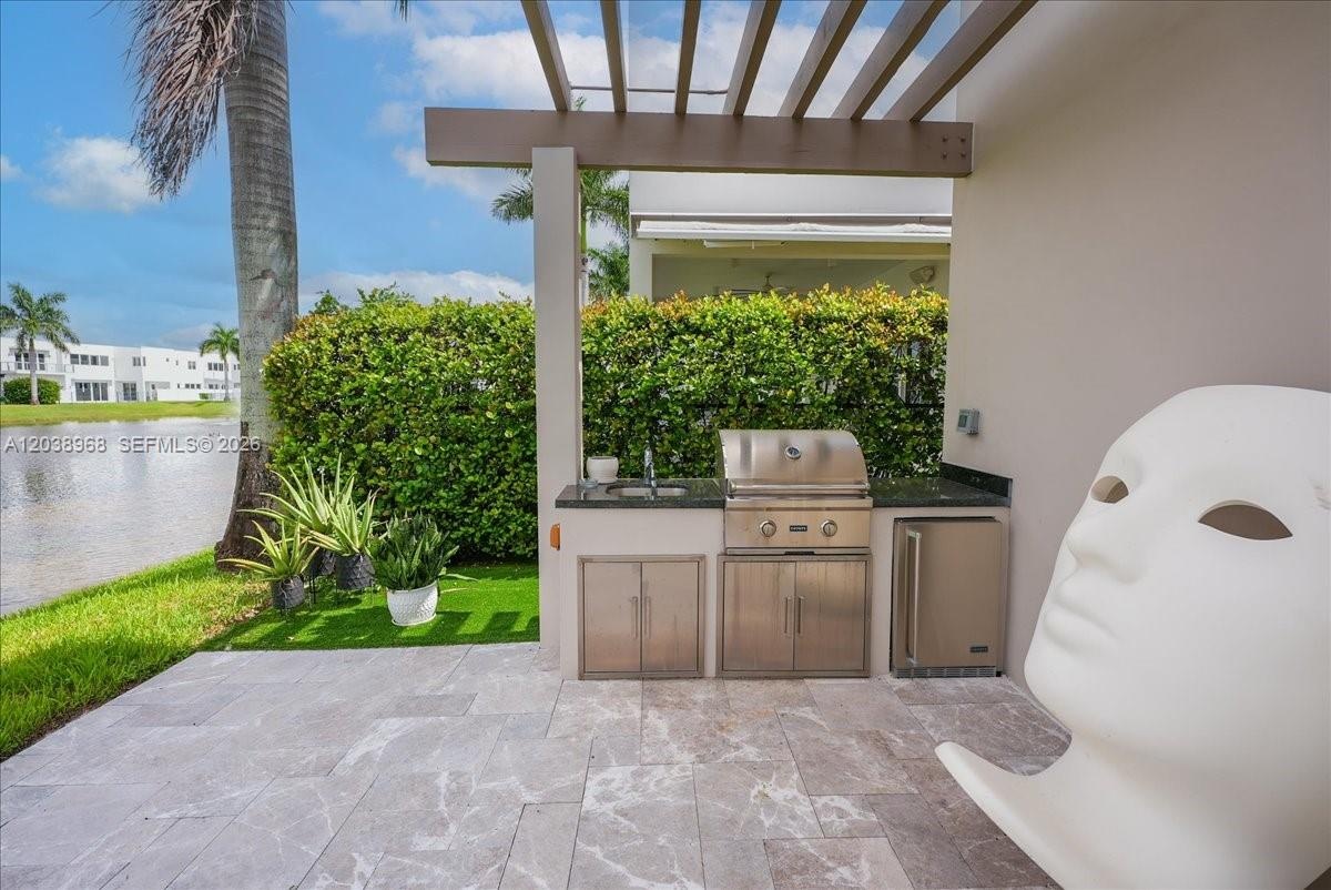 Outdoor kitchen featuring a stone countertop, built-in stainless steel grill, under-counter refrigeration, and sink, situated under a wood-beam pergol