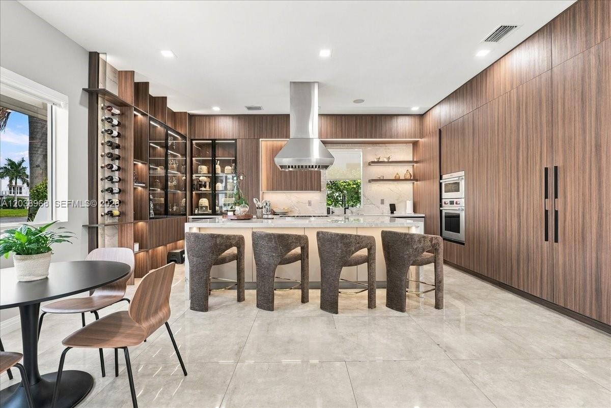 Modern kitchen featuring full-height wood-finish cabinetry, a large central island with stone surfacing, stainless steel range hood, integrated wall o