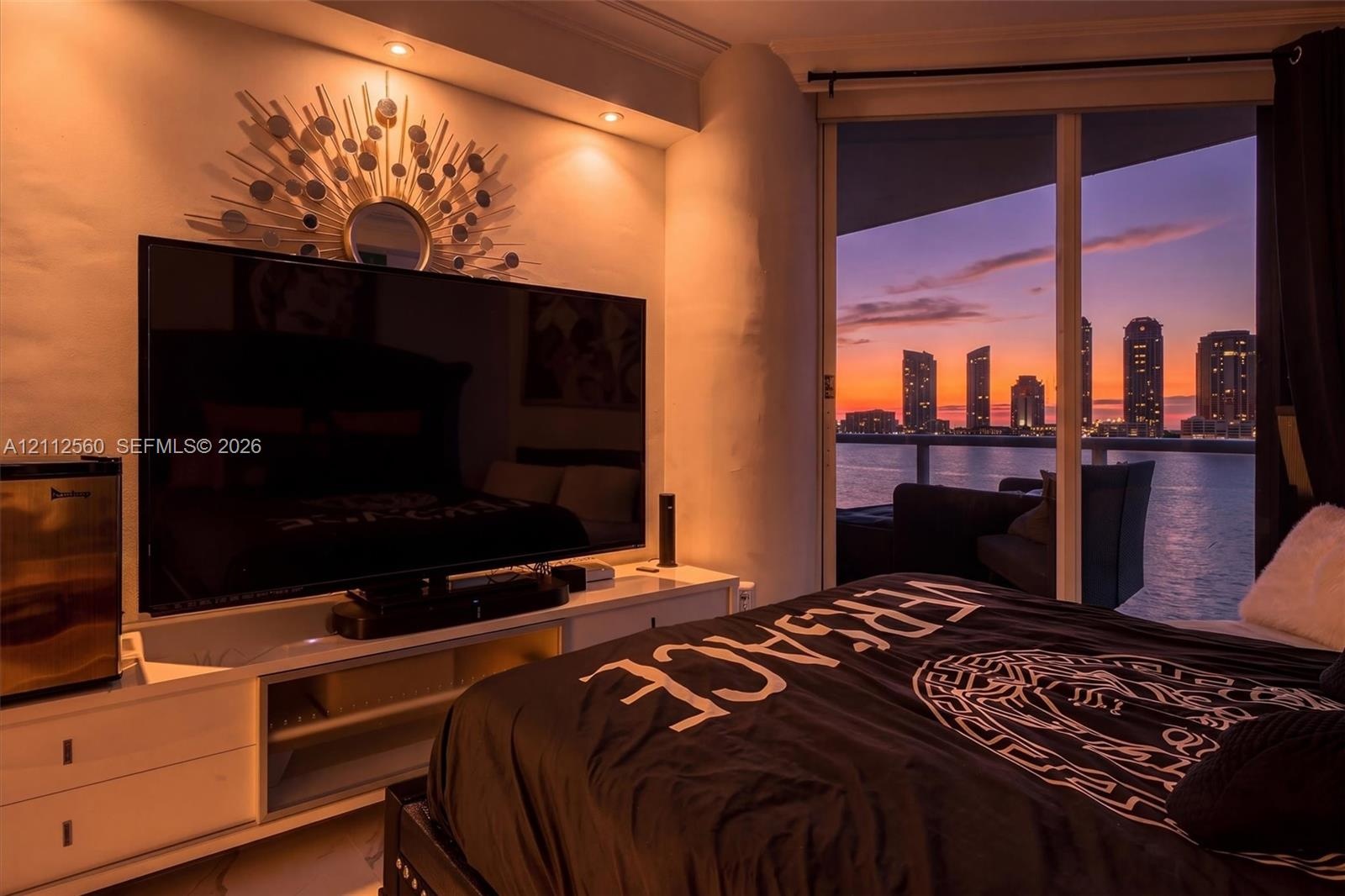 Floor-to-ceiling glass sliding doors, built-in recessed ceiling lighting, and private balcony with metal railing, set against a waterfront city skylin