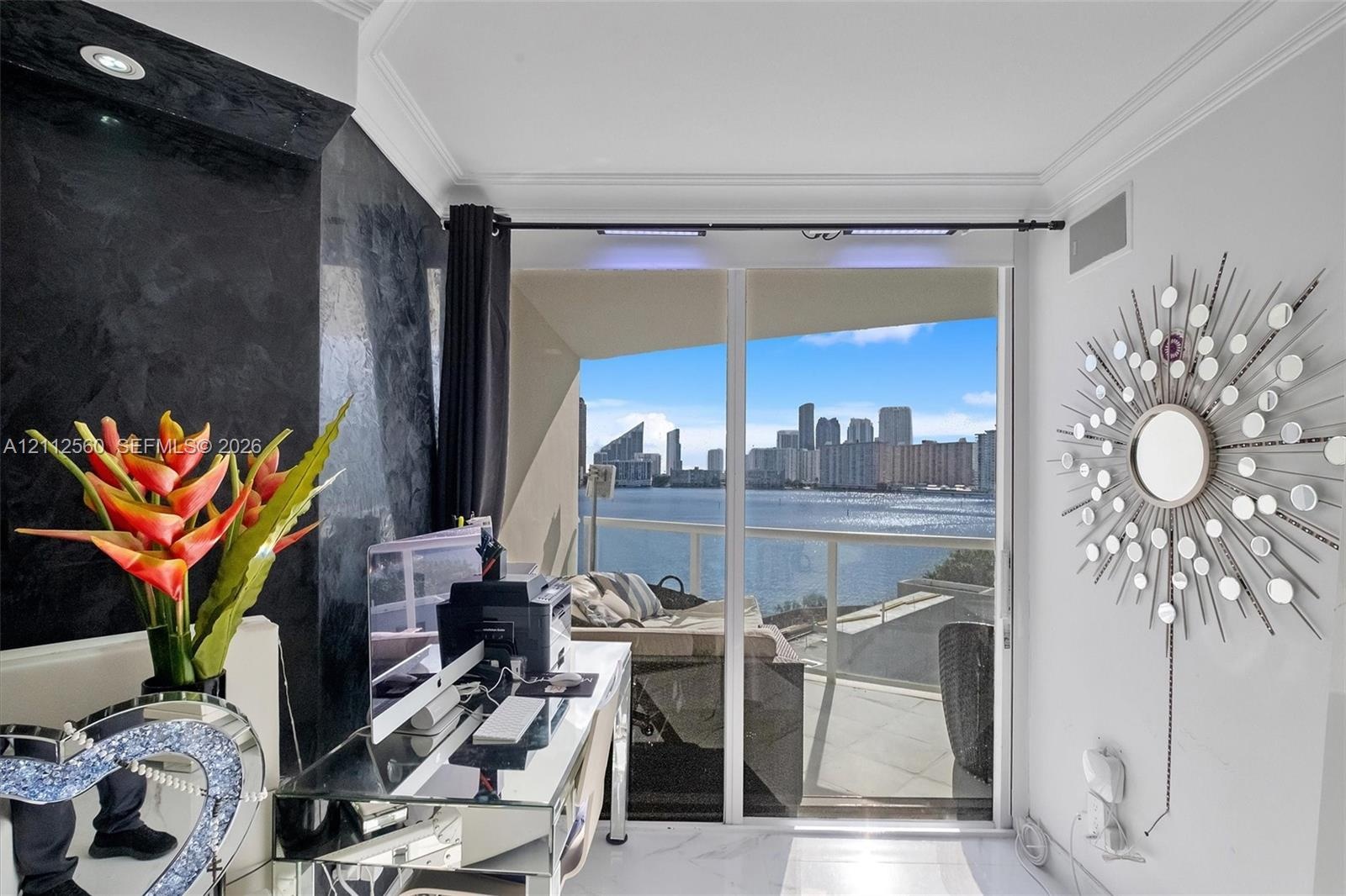 Floor-to-ceiling glass sliding doors opening to a balcony with water and skyline scenery, featuring a textured black feature wall, recessed lighting, 