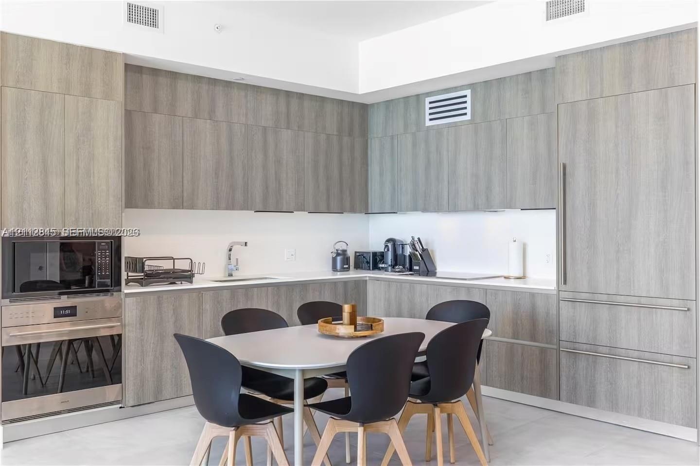 Kitchen featuring flat-panel wood-finish cabinetry, integrated refrigerator, built-in stainless steel microwave and wall oven, and light-toned solid s