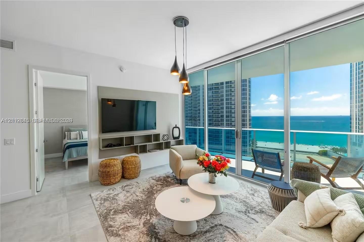 Floor-to-ceiling glass wall with ocean backdrop, private balcony, neutral large-format tile flooring, built-in wall-mounted media console, and triple-