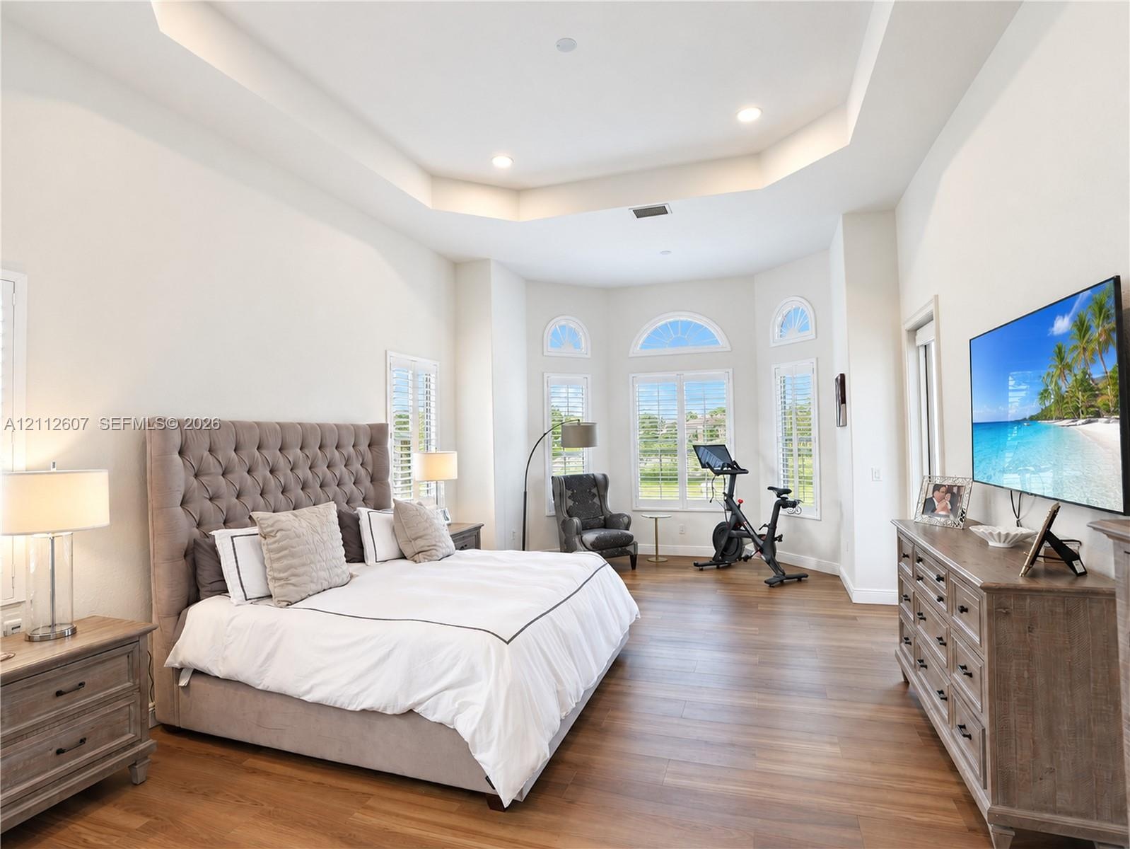 Tray ceiling with recessed lighting, wood-finish flooring, and expansive bay windows featuring arched transoms and plantation shutters