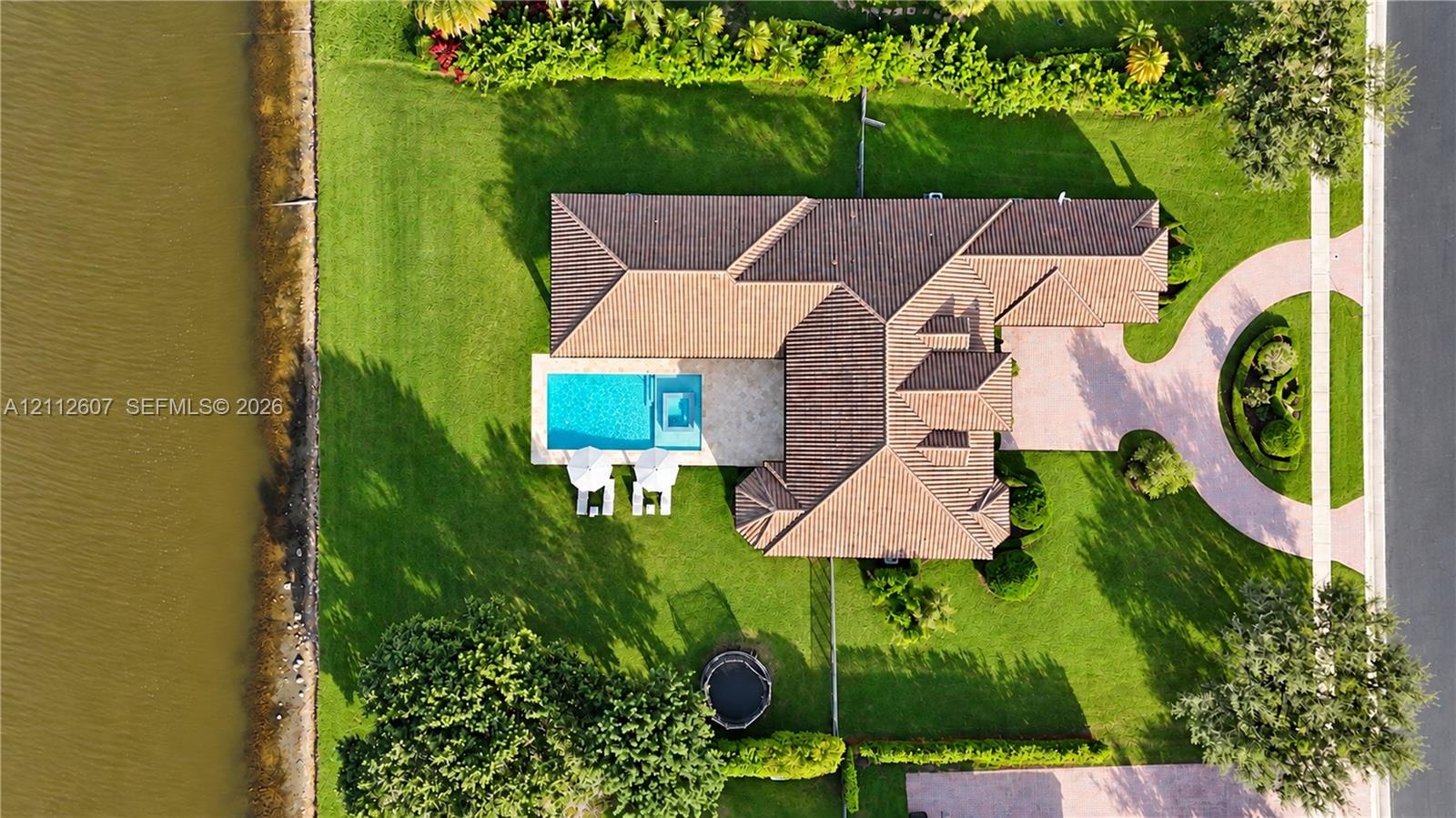 Aerial perspective of a waterfront property featuring a tile roof, an in-ground swimming pool with an integrated spa, and paved circular driveway