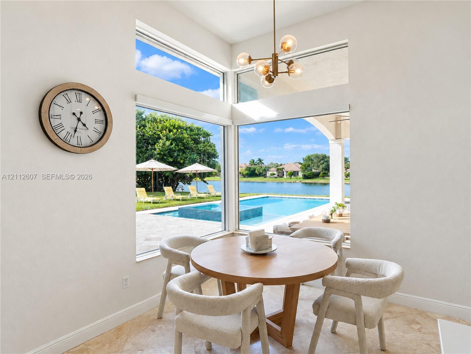 High-ceiling dining area featuring expansive corner windows, a multi-globe metal chandelier, and stone-finish flooring with outdoor water and pool acc