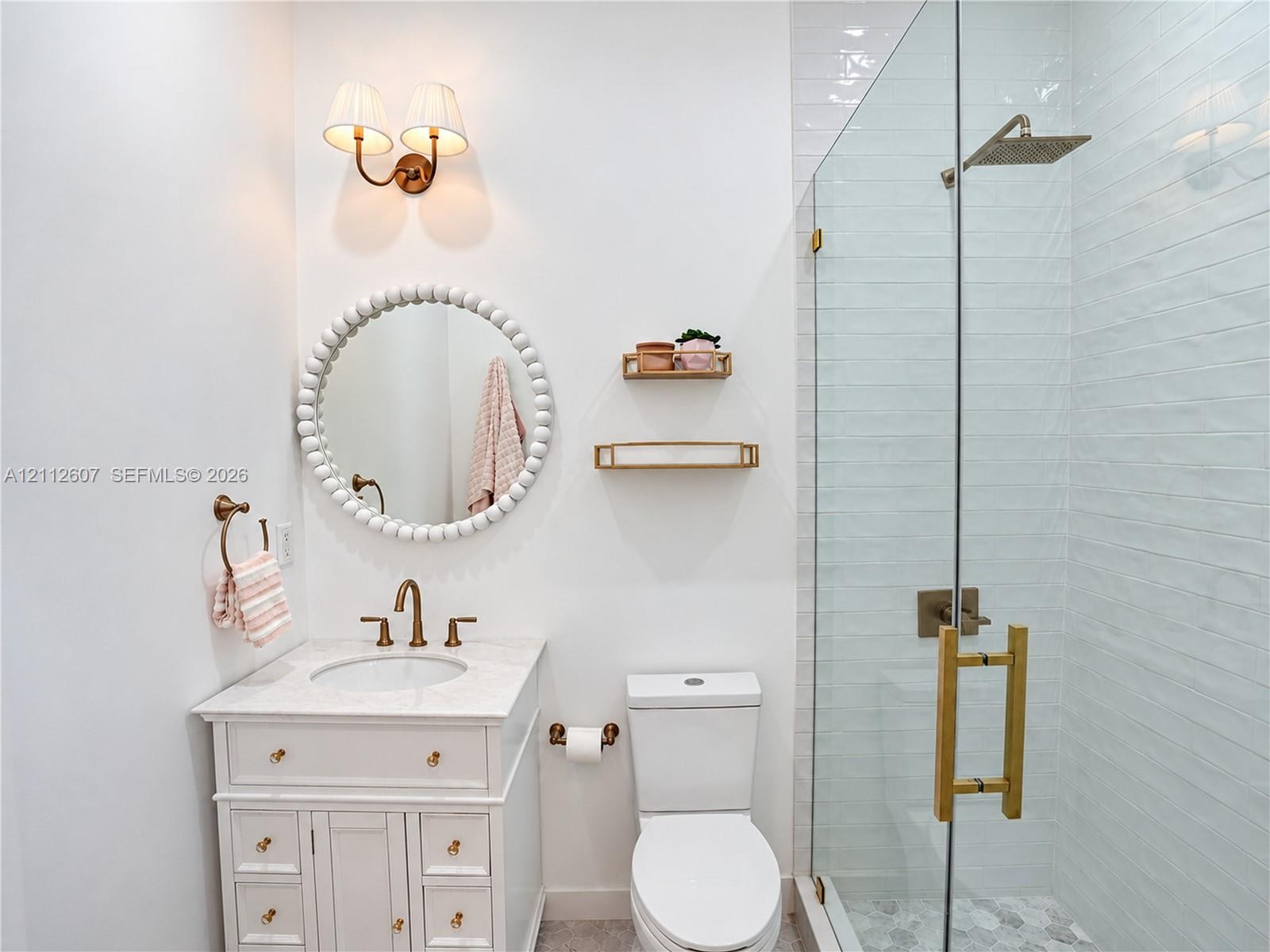White vanity cabinet with a stone countertop, wall-mounted dual-bulb light fixture, circular bead-detail mirror, and a glass-enclosed shower with a ra